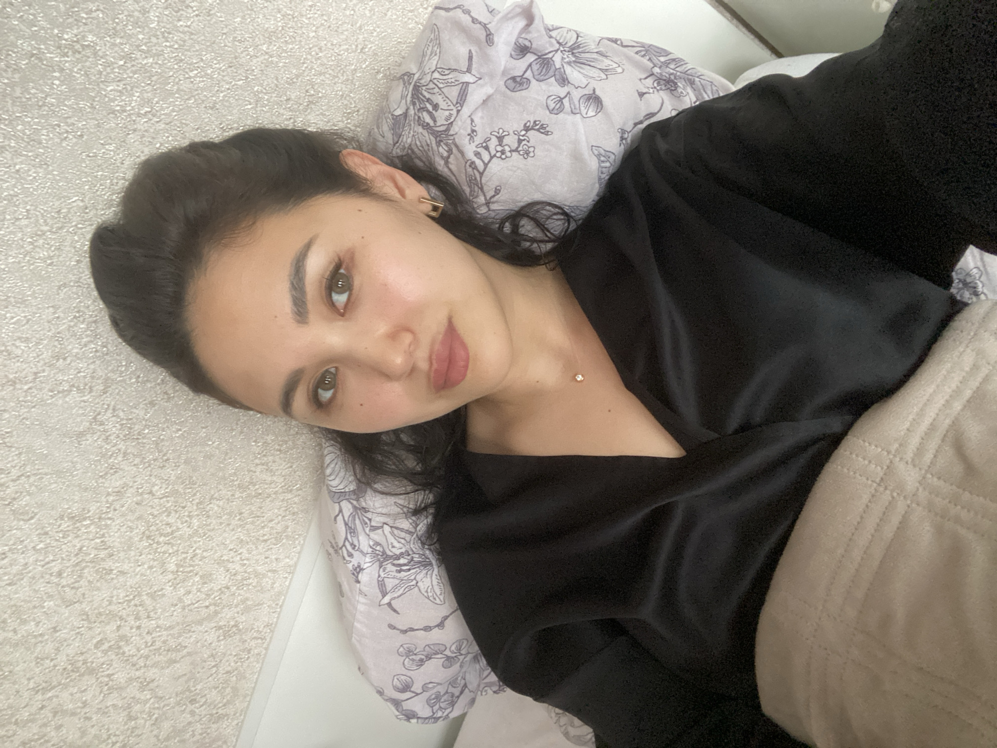 A woman lies on a bed covered with patterned bedding. She wears a black robe and a necklace. A tan blanket is pulled up to her chest. The location is not identifiable as Vysokoye, Russia based on the provided image.
