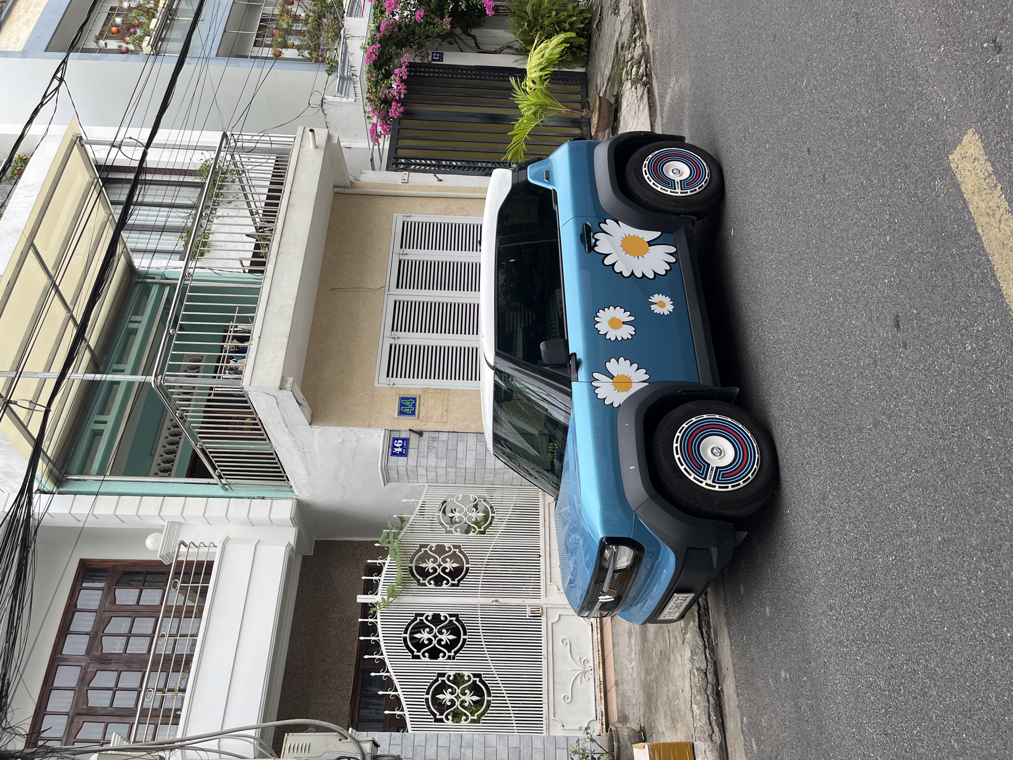The image shows a blue electric car with white daisy stickers parked on a street in Nha Trang, Vietnam. The car is in the foreground, facing right. Behind the car is a white building with balconies and shuttered windows. A white ornate gate is visible to the left of the car, and a darker metal gate with pink flowers is to the right. The street is asphalt, with a yellow line painted on it. The sky is overcast. There are no people visible in the image. The license plate of the car reads "79A-564.22". The building has house numbers "46" and "44" visible on its facade.
