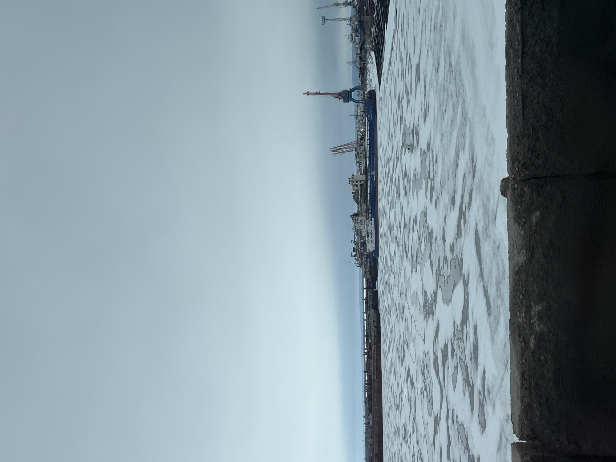 The image shows a view of the Kama River waterfront in Chaikovsky, Russia. Ice floes cover the water surface, and along the shore, a concrete embankment is visible. In the background, a harbor with cargo ships and cranes can be seen. The sky is overcast and grey.