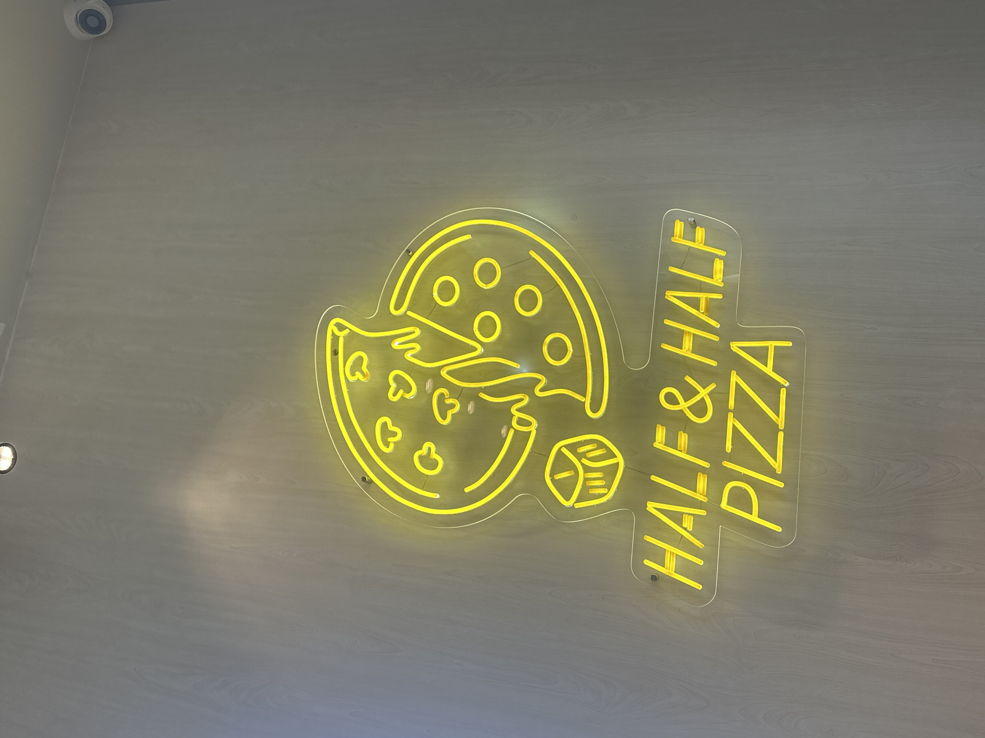 A neon yellow sign reading "HALF & HALF PIZZA" is mounted on a light-colored, wood-paneled wall. The sign features a graphic of a pizza, with one half depicted with mushroom toppings and the other with cheese circles. A small, cube-shaped object is also part of the graphic, positioned to the left of the pizza. A security camera is visible in the upper right corner of the image. The location is identified as Gyumri, Armenia.