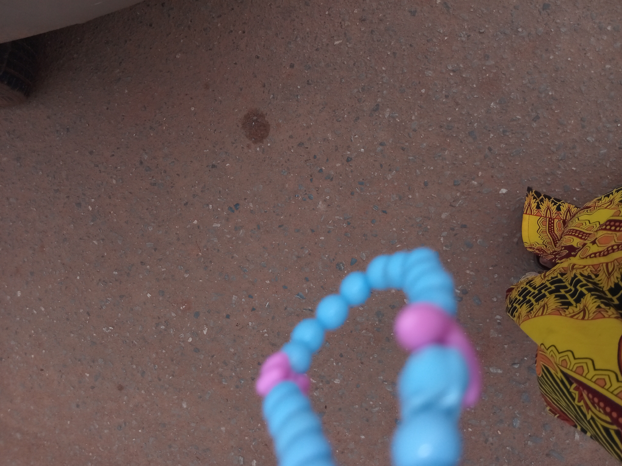 The image shows a ground-level view of an outdoor area, possibly in Shagamu, Nigeria, though there are no specific landmarks that definitively identify the location. The primary surface is reddish-brown, speckled asphalt or concrete. In the lower right corner is a portion of yellow and black patterned fabric. In the foreground, a blue and pink plastic toy made of segmented beads forming a curve. The tire of a vehicle is visible on the left side of the frame. There is a single, small, dark stain on the ground.