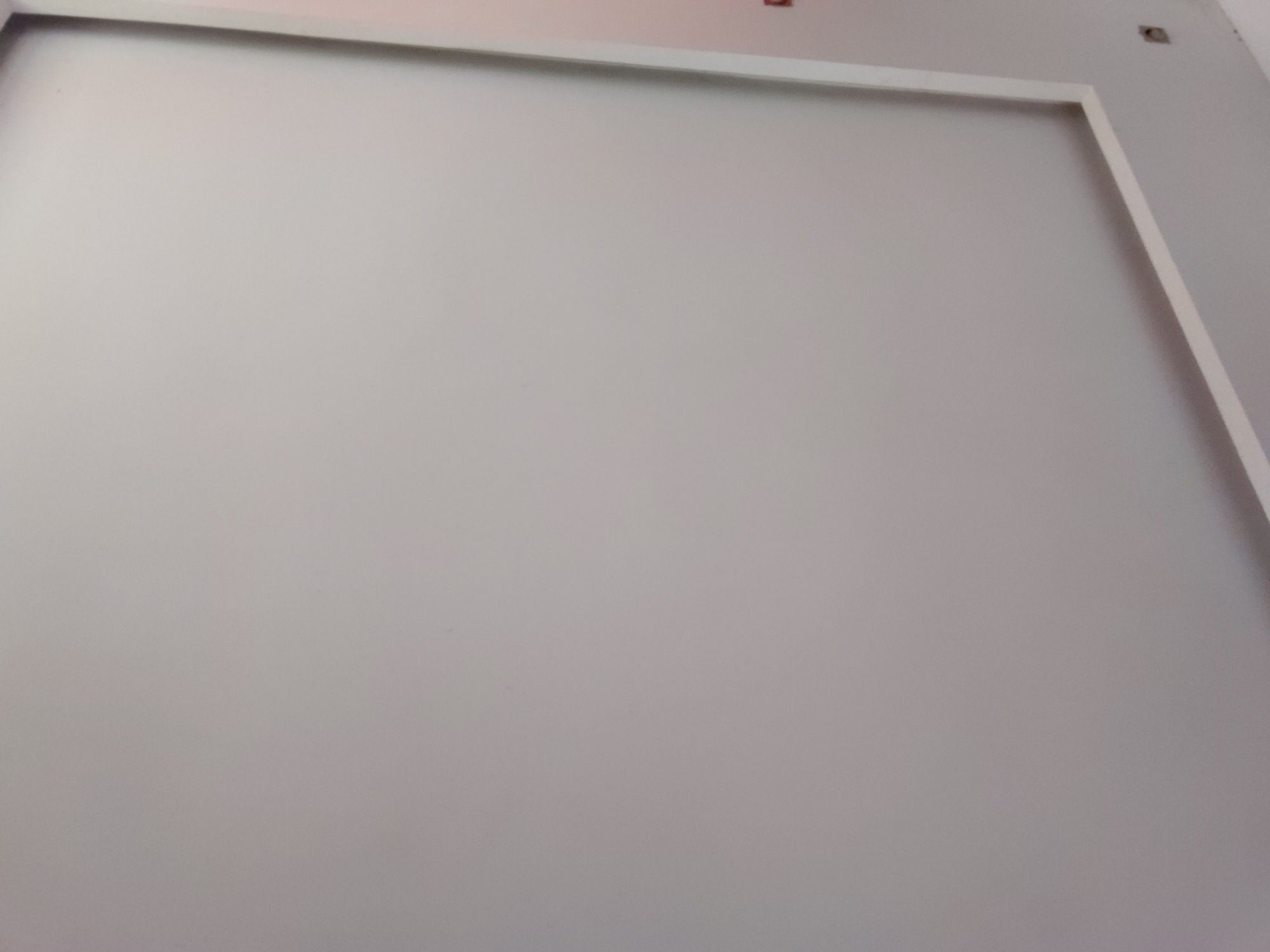The image displays a plain, light grey or off-white wall, likely indoors. A prominent white trim or molding frames a large rectangular section of the wall, visible along the top and right edges. This trim creates a distinct, clean border for the central wall area, which is uniformly colored. In the upper right portion of the wall, beyond the white frame, two small, dark, square or rectangular openings are visible, which could be electrical outlets, light switch cutouts, or mounting points.
No people are present, and no specific activities or events are taking place; the scene is static, focusing purely on the architectural detail of the wall. The lighting appears even and diffused, consistent with an interior setting. There are no notable location cues within the image itself to identify Santa Ana, Venezuela, other than the provided context. No text is visible anywhere in the frame. The overall impression is one of a modern, minimalist, and possibly unfinished or newly painted room.