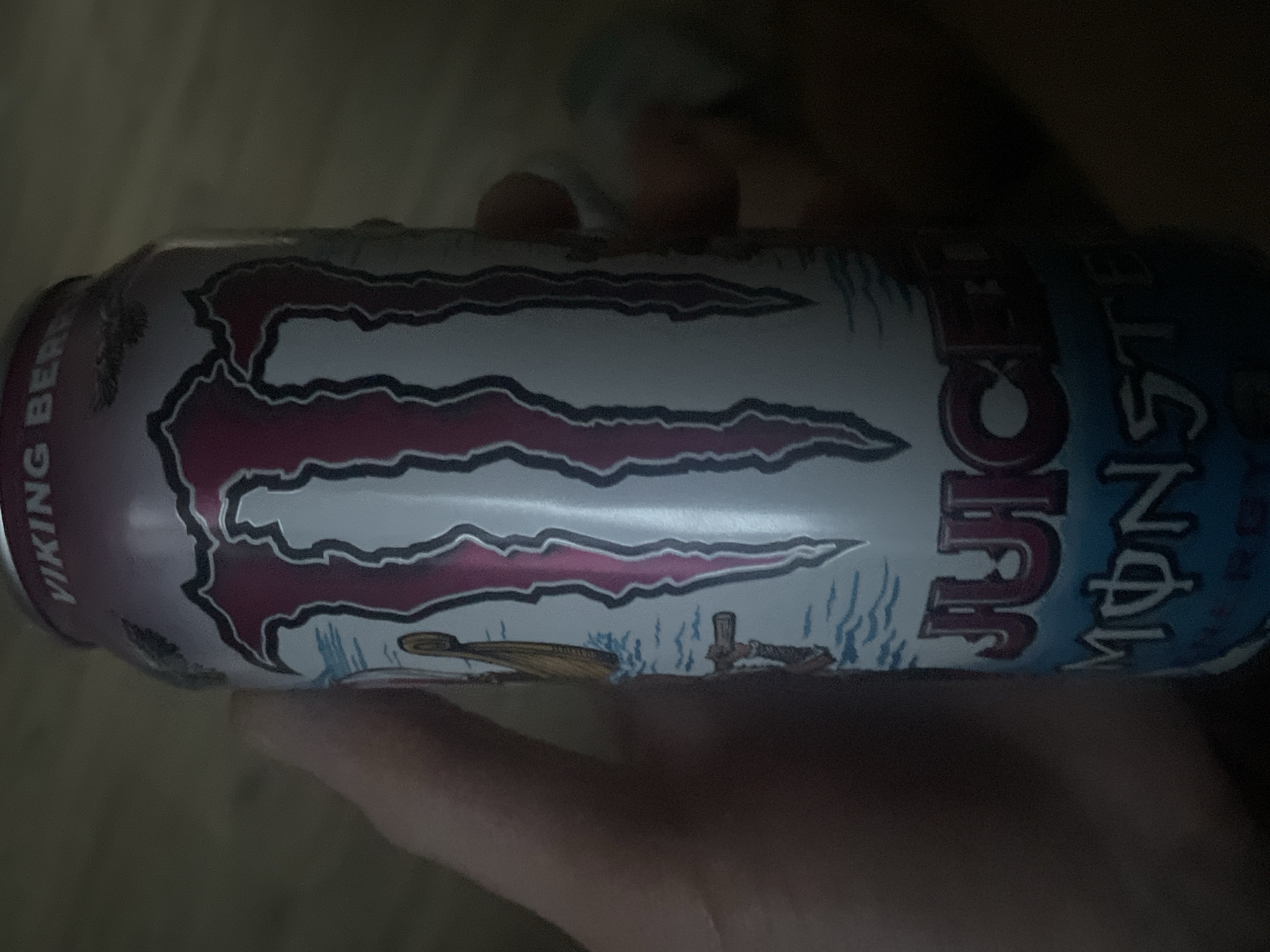 A hand is holding a can of Monster Energy drink, specifically the "Juice Monster Viking Berries" flavor. The can is primarily white with pink and dark pink accents. The prominent Monster logo is visible, along with text that reads "JUICE" and "MONSTER". The background is dark and out of focus, making it impossible to discern any specific details of the environment, including the city of Paczkowo, Poland. No people other than the implied hand holding the can are visible.