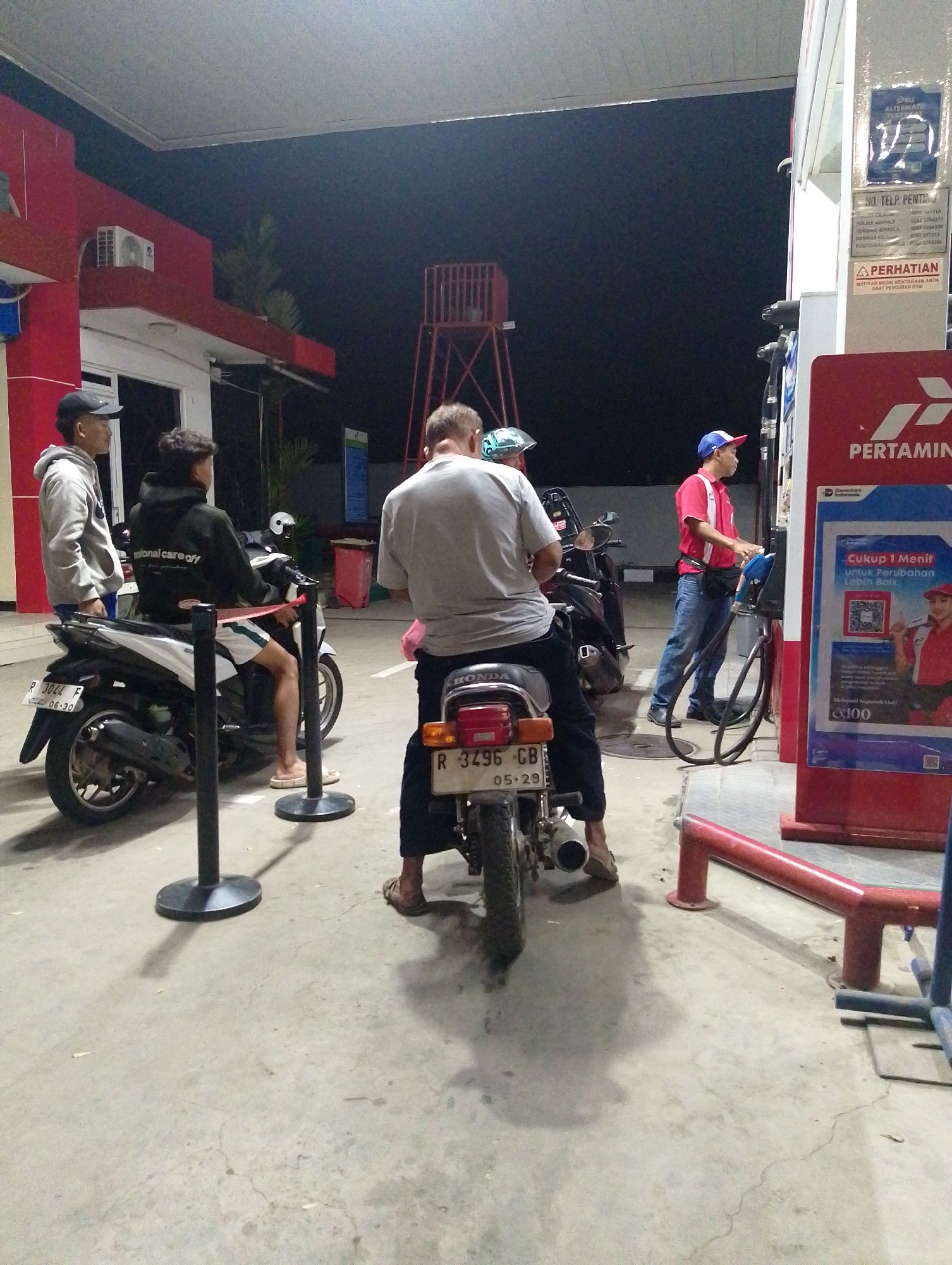 The image shows a night scene at a Pertamina gas station in Adipala, Indonesia. In the foreground, a person on a motorcycle with license plate number R 3044 F is being refueled by a gas attendant. The attendant, wearing a red uniform and blue cap, is operating the fuel pump. To the left, two other individuals stand near a motorcycle, one wearing a gray hoodie and a black cap, the other a black hoodie. Another motorcycle with license plate number R 3496 GB is parked to the right. The gas station features red and white structures, a red water tower in the background, and various signage. The ground is paved concrete.
