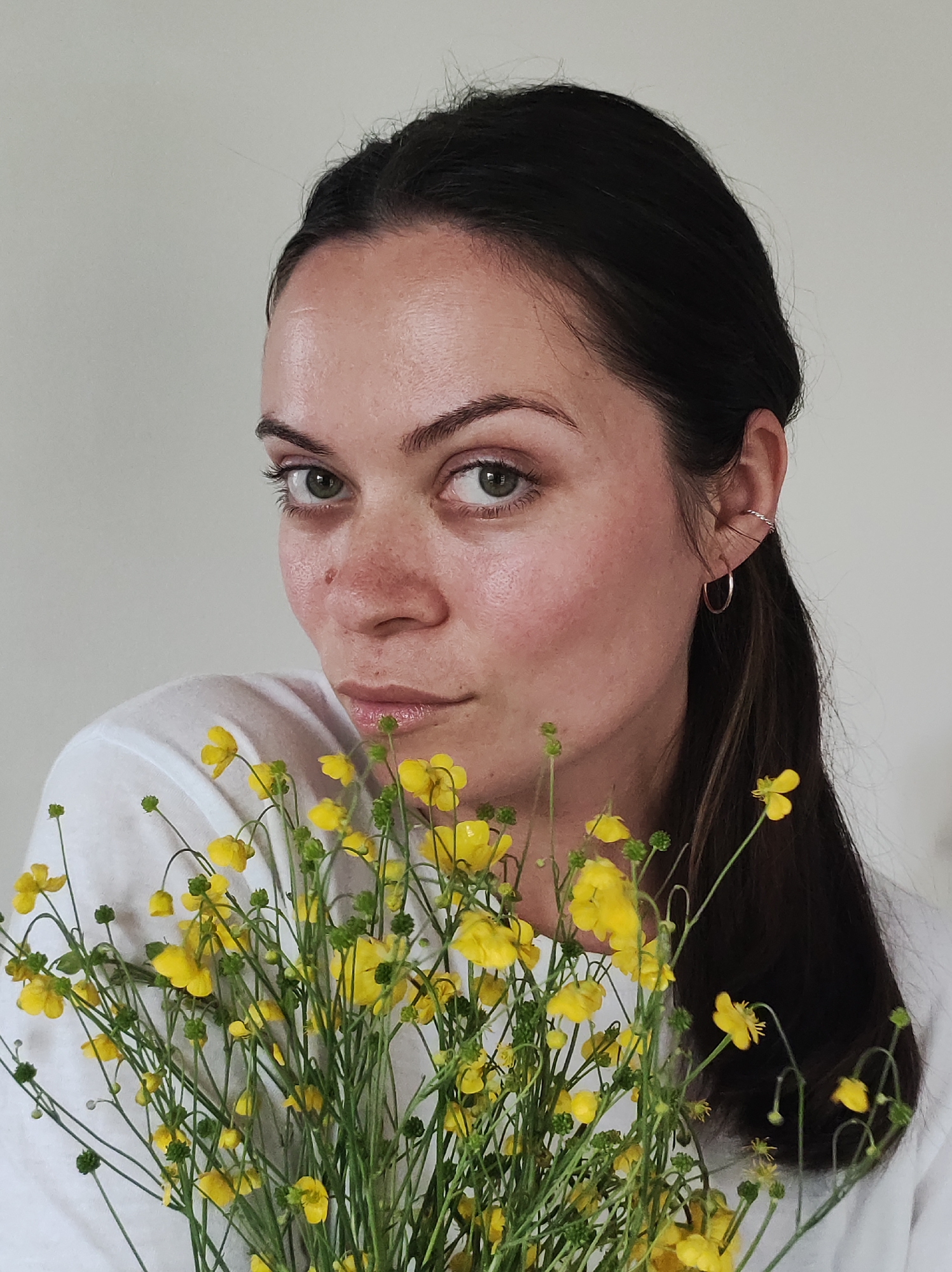 A close-up portrait features a woman with dark hair and striking green eyes, looking directly at the viewer. Her dark hair is pulled back, revealing a natural complexion with visible freckles scattered across her nose and cheeks, and slight redness around the nasal area. Her gaze is direct and intense, with a subtly serious or contemplative expression on her lips.
She is wearing a simple white t-shirt. On her visible right ear (viewer's left), she sports two pieces of jewelry: a delicate gold-toned hoop earring in her lobe and a silver-toned, twisted ear cuff positioned higher on her ear.
She holds a vibrant bouquet of small, yellow flowers, resembling buttercups or wild buttercups, in front of her lower face and upper body. The bright yellow petals and green stems provide a splash of color against her white shirt. The background is a plain, light-colored wall, indicating an indoor setting with soft, even lighting. No text is visible in the image. The overall aesthetic is natural and intimate. This image is potentially taken in Foky, Russia.