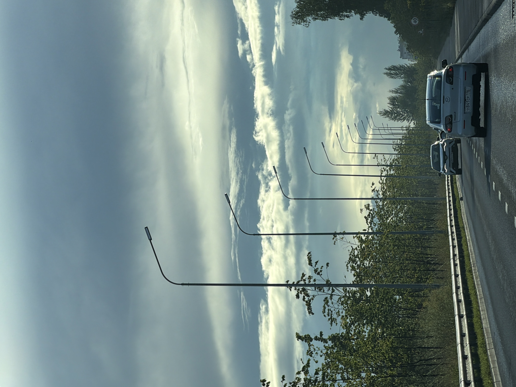 The image depicts a view from a car traveling on a highway in Kazan, Russia. The sky is filled with dramatic, cloudy formations, with patches of sunlight breaking through. A line of streetlights extends along the side of the road, interspersed with trees and vegetation. Several cars are visible on the highway, traveling in the same direction. A speed limit sign indicating 40 is visible on the right side of the road. The overall atmosphere suggests overcast weather conditions.