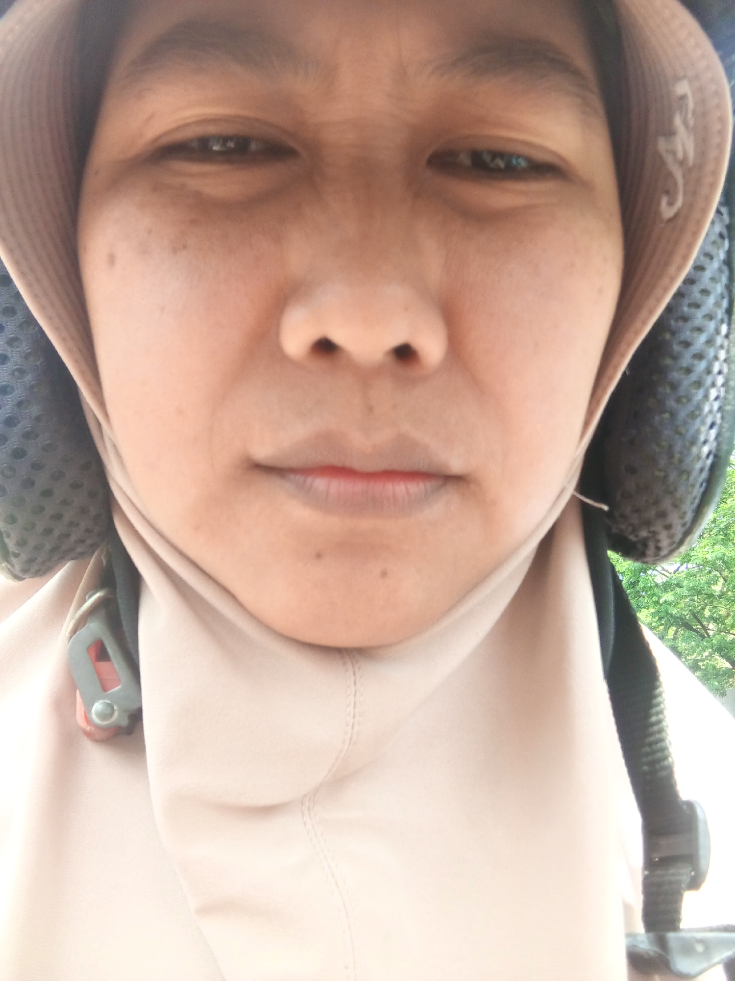 A close-up, low-angle, portrait photograph captures the face of a person wearing a light brown head covering and a helmet. The individual's skin has a warm tone with visible pores and freckles. The eyes are partially visible and appear dark. The mouth is closed, with the upper lip slightly fuller than the lower. The helmet, which is dark and textured, encircles the head, with a strap and buckle visible on the left side of the frame. The background consists of out-of-focus green foliage, suggesting an outdoor environment. The image appears to have been taken in Banyudono, Indonesia.