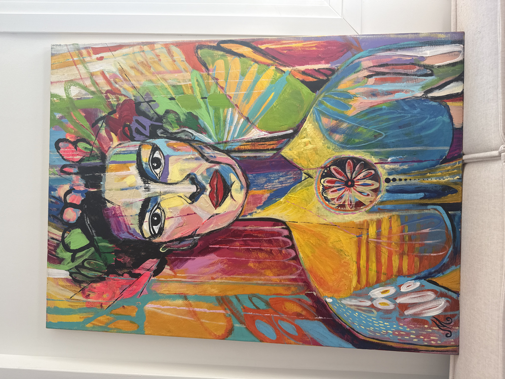This is a photograph of a colorful, abstract painting on canvas, depicting a woman's portrait. The painting is vibrant, with bold brushstrokes and a mix of warm and cool colors, including yellow, orange, pink, blue, green, and purple. The woman has dark hair adorned with colorful elements, possibly flowers or leaves, and her face is rendered with sharp lines and varied hues. She is wearing a yellow garment with a circular, floral-like motif at the chest. The background is a chaotic yet harmonious blend of abstract shapes and colors, suggesting a tropical or natural setting. The painting is leaning against a white wall, with a sliver of a white window frame visible on the right. The bottom of the painting rests on a light-colored, textured surface, possibly a sofa or cushion. The signature "M" in black cursive is visible on the lower left of the painting. The photograph was taken indoors. The city of Neoi Epivates, Greece is not visible in this image.