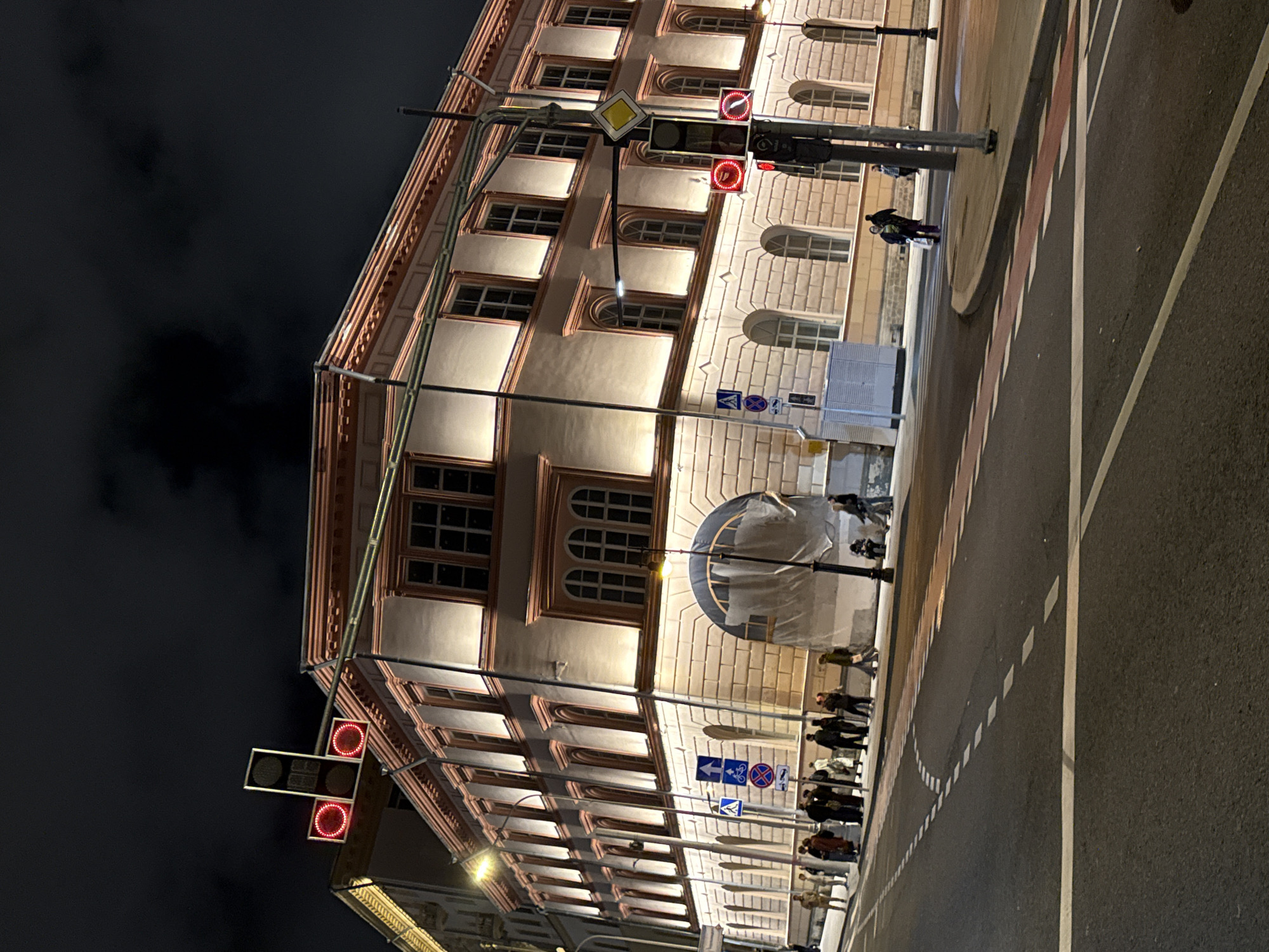 The image shows a nighttime street scene in Moscow, Russia. A large, ornate building with illuminated windows and decorative trim is prominent on the right side of the frame. The building appears to be made of light-colored stone with a reddish-brown decorative band along the roofline and around some of the windows.
In the foreground, a street with lane markings is visible. Several traffic lights are present, with red lights illuminated on at least two of them. Pedestrian crossing markings are also visible on the road.
On the sidewalk in front of the building, a group of people are standing and walking. Some are dressed in dark clothing, while others wear lighter colors. There are also street signs and a lamppost. The sky is dark and cloudy, indicating nighttime. The overall atmosphere is urban and somewhat subdued due to the low light conditions.
