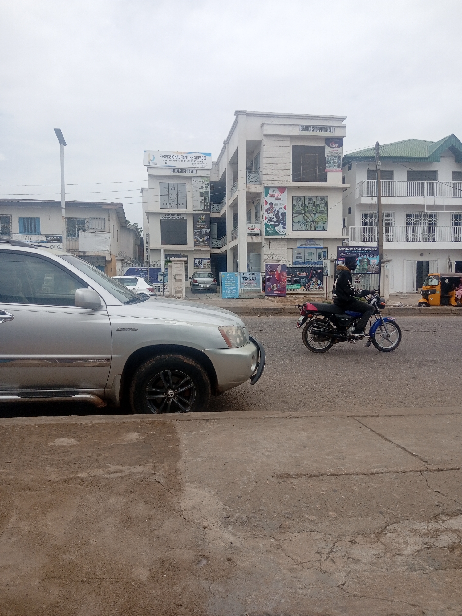In Sardauna, Nigeria, on a cloudy day, a silver Toyota Highlander SUV is parked on the left side of the frame, facing right, with its front half visible. The vehicle's tires and part of its side are in focus. Beyond the car, a paved road stretches into the distance. On the right side of the road, a man wearing a dark helmet and jacket rides a blue and black motorcycle, also facing right. Behind him, a yellow auto-rickshaw with three wheels is parked. In the background, several multi-story buildings line the street. The building directly behind the motorcycle features prominent signage including "IBRAHKA SHOPPING MALL 1" and "PROFESSIONAL PRINTING SERVICES." Other buildings show various advertisements and architectural details, including balconies with white railings and windows. The environment appears to be an urban commercial area.