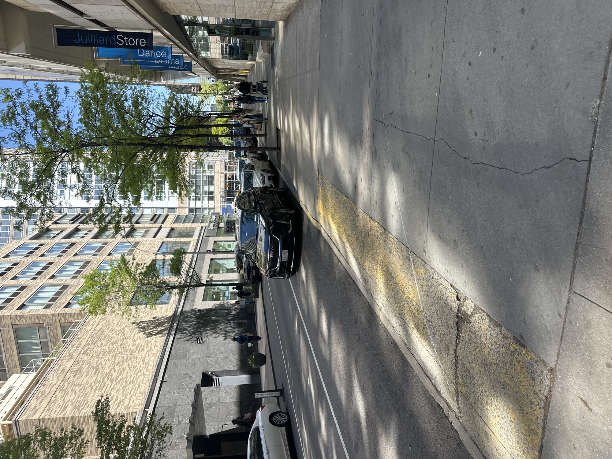 The image depicts a city street scene, likely in New York City, United States, based on the "Juilliard Store" and "Dance" signs visible in the upper left. The perspective is from the sidewalk looking towards the street. A row of parked cars lines the left side of the street, with a black SUV prominently featured in the foreground. Several pedestrians are walking along the sidewalk in the distance. Buildings of varying heights and architectural styles are visible in the background, interspersed with trees that cast shadows on the street. The sidewalk is made of concrete and shows signs of wear and cracks. A curb painted with yellow markings separates the sidewalk from the street. The lighting suggests it is daytime with bright sunlight. A "No Parking" sign is visible near a white car parked further down the street.