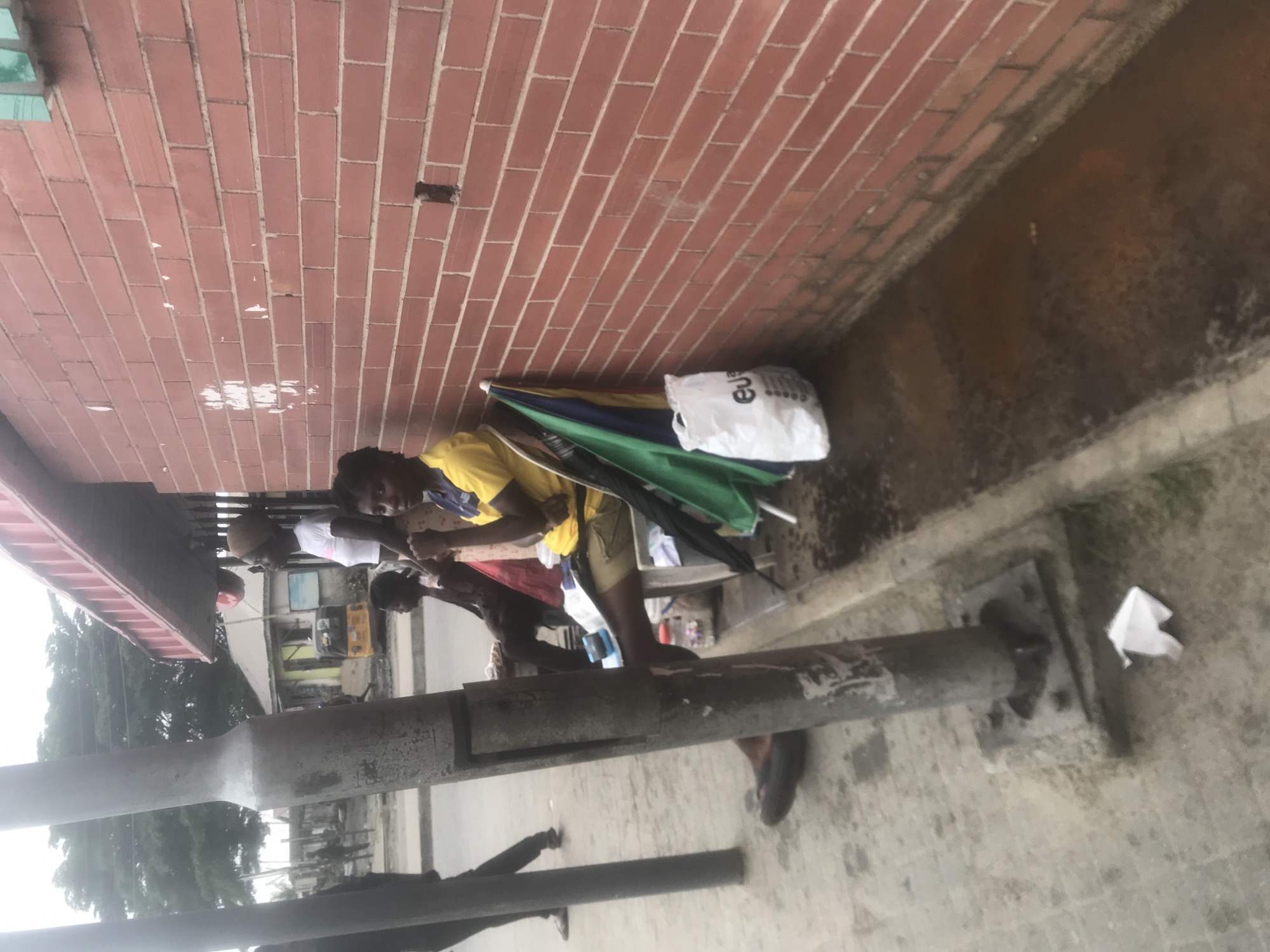 The media file shows a group of people standing near a brick wall and a metal pole. The people appear to be gathered under an overhang or awning, possibly seeking shelter. There are several objects present, including an umbrella with colorful sections, a white shopping bag with some text on it, and some clothing. The ground is paved with concrete.
The scene appears to be outdoors, with trees and buildings visible in the background, suggesting an urban or semi-urban environment. The lighting suggests it is daytime, though the weather is unclear. The people's expressions are not clearly discernible due to the image quality and orientation.
One person in the foreground is wearing a yellow shirt and khaki shorts. Another person is wearing a white t-shirt. There are other individuals partially visible. The overall activity seems to be people congregating in a public space.
Notable details include the texture of the brick wall and the weathered appearance of the metal pole. There is a white plastic bag near the umbrella, with visible text that appears to be part of a logo or brand name. The image is oriented vertically, making it appear as if the scene is sideways.