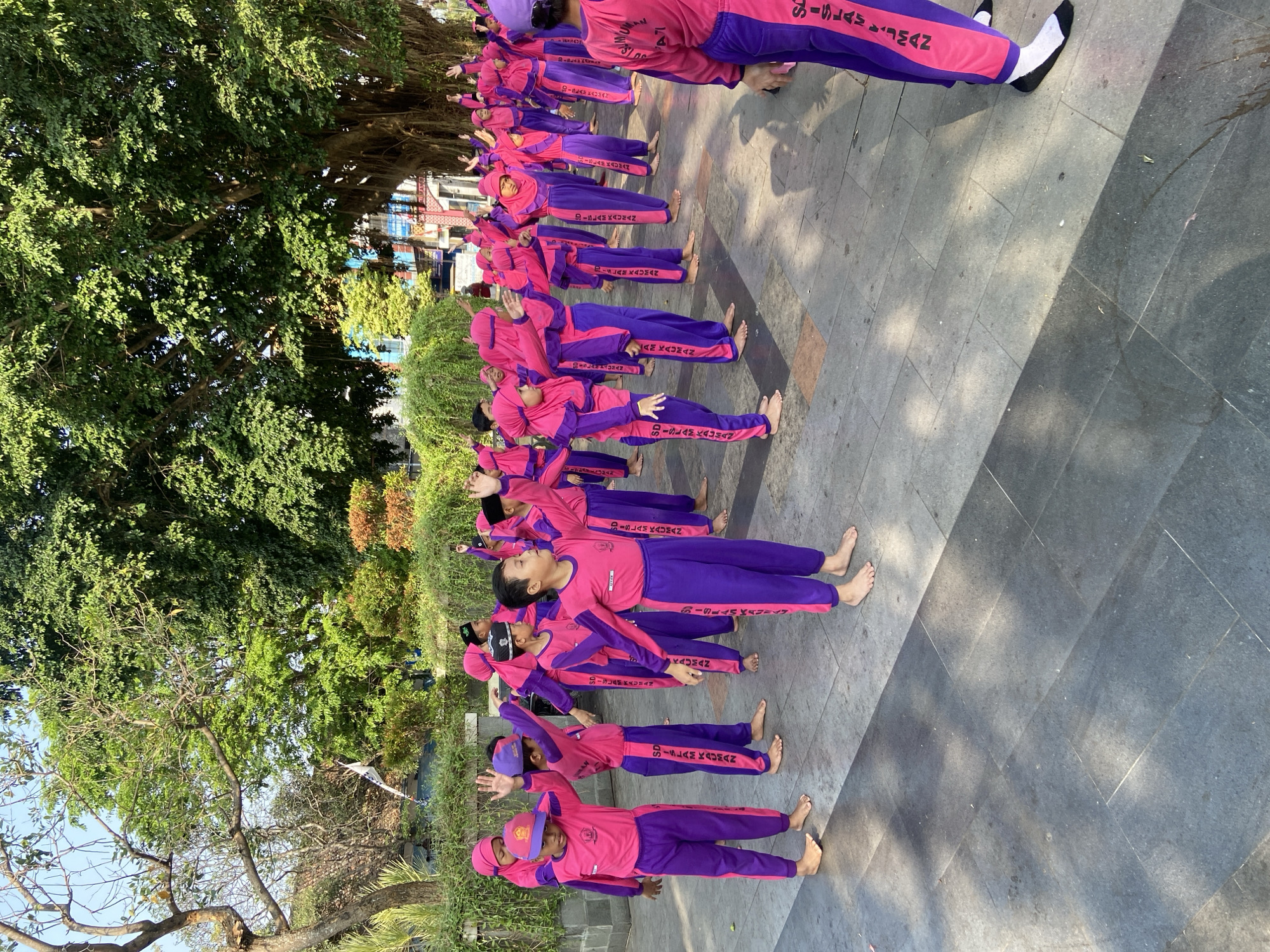A group of children, likely students, are gathered outdoors in what appears to be a public plaza in Kecamatan Pati, Indonesia. They are all wearing matching pink and purple uniforms, consisting of long-sleeved shirts and pants. Most of the children have their right arms raised and bent at the elbow, with their palms facing forward. Some are barefoot, while others are wearing socks. The children are arranged in several rows, facing the same direction. Behind them, a large, mature tree with dense foliage dominates the background. The ground is paved with large, grey stone tiles. The overall impression is of an organized group activity, possibly a school event or physical education class, taking place in a park-like setting within the city.