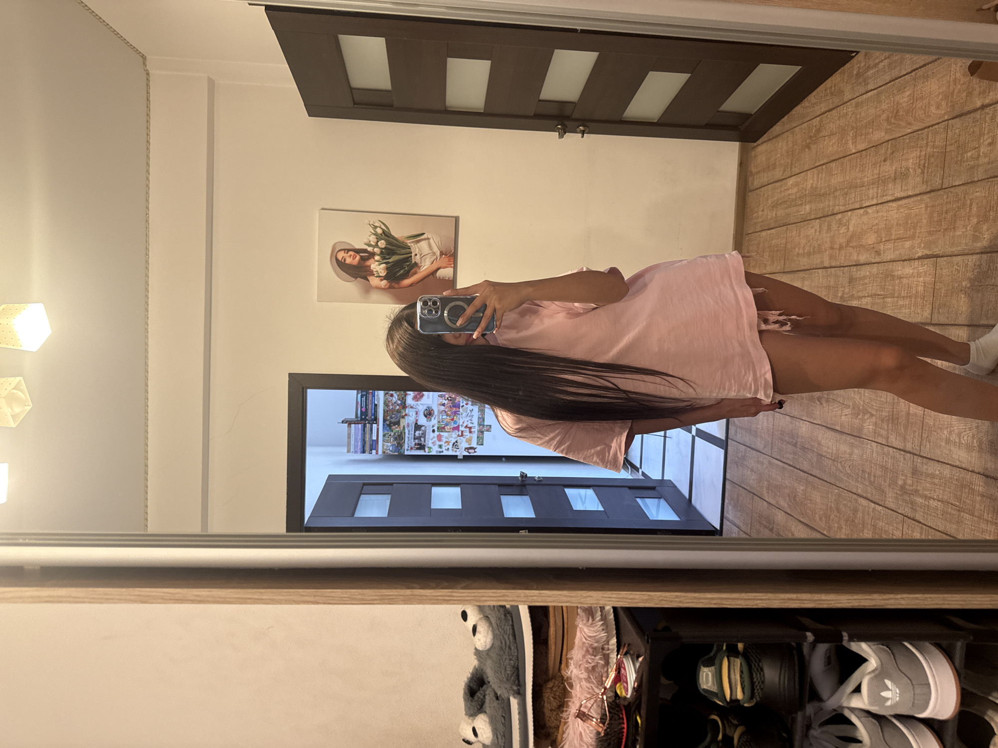 A young woman is standing in front of a mirror, taking a selfie. She has long, dark hair and is wearing a pink t-shirt and white socks. She is in a room with wooden floors and a dark brown door with glass panels. To the left of the mirror is a shoe rack with several pairs of shoes, including a pair of grey Adidas sneakers. On the wall to the right of the woman is a framed picture of a woman wearing a white hat and holding a bouquet of tulips. The room appears to be an entryway or hallway, possibly in an apartment in Chișinău, Moldova.