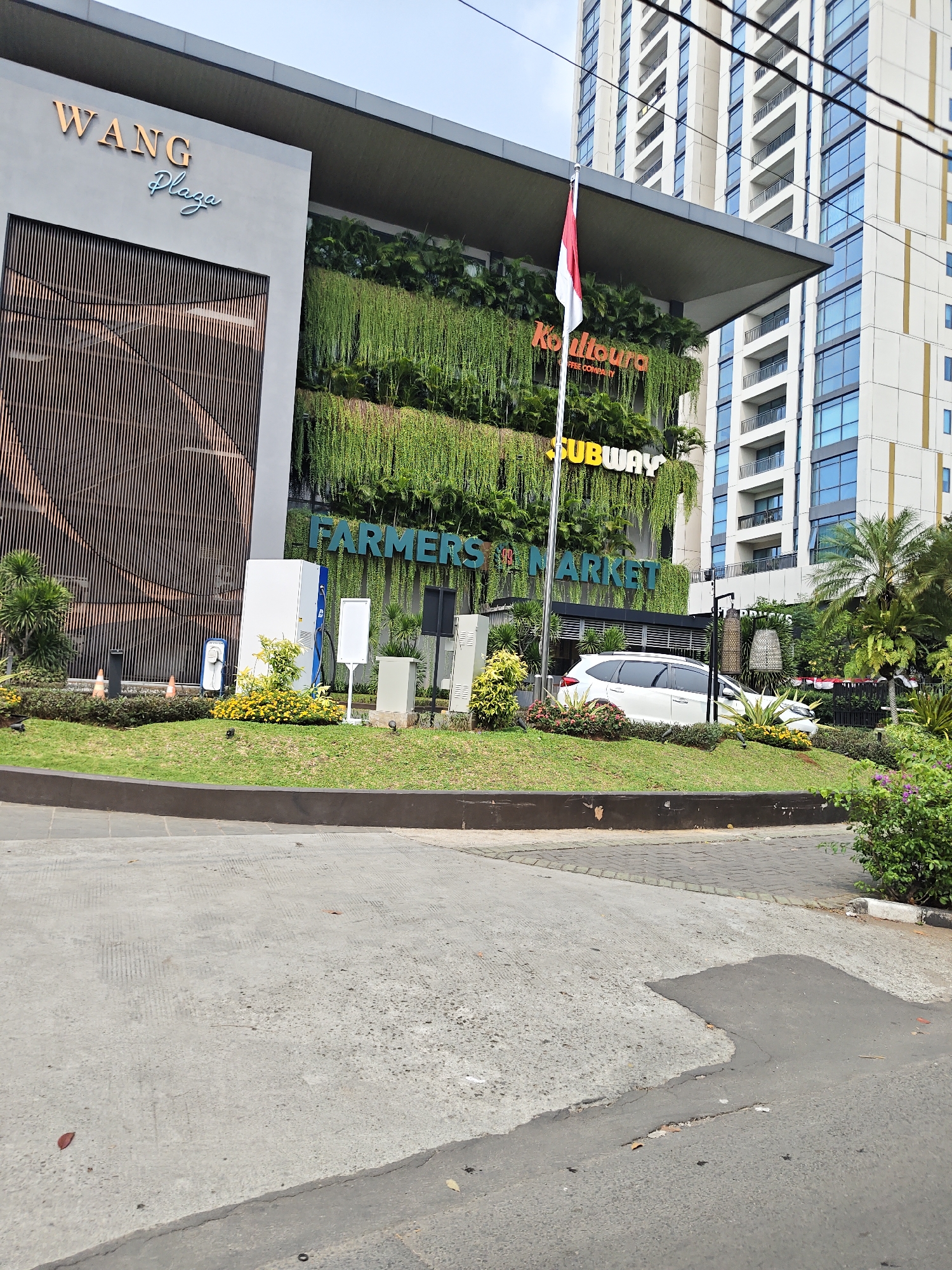 The image shows a modern building exterior with the sign "WANG Plaza" and "Plaza" in script font. Below the plaza sign, there is a vertical slatted wooden panel on the building's facade. The building features a vertical garden covering a large section of its exterior. Signs for "KopiToura", "Subway", and "FARMERS MARKET" are visible on the building. A red and white Indonesian flag flies from a flagpole. A white SUV is parked on a grassy slope in front of the building, with various plants and landscaping. Several utility boxes and what appear to be charging stations are located on the grass. In the background, a tall, modern residential or office building with many windows is visible. The foreground shows a concrete road and walkway. The scene is set in a city environment, consistent with urban development in West Jakarta, Indonesia. There are no people visible in the image.