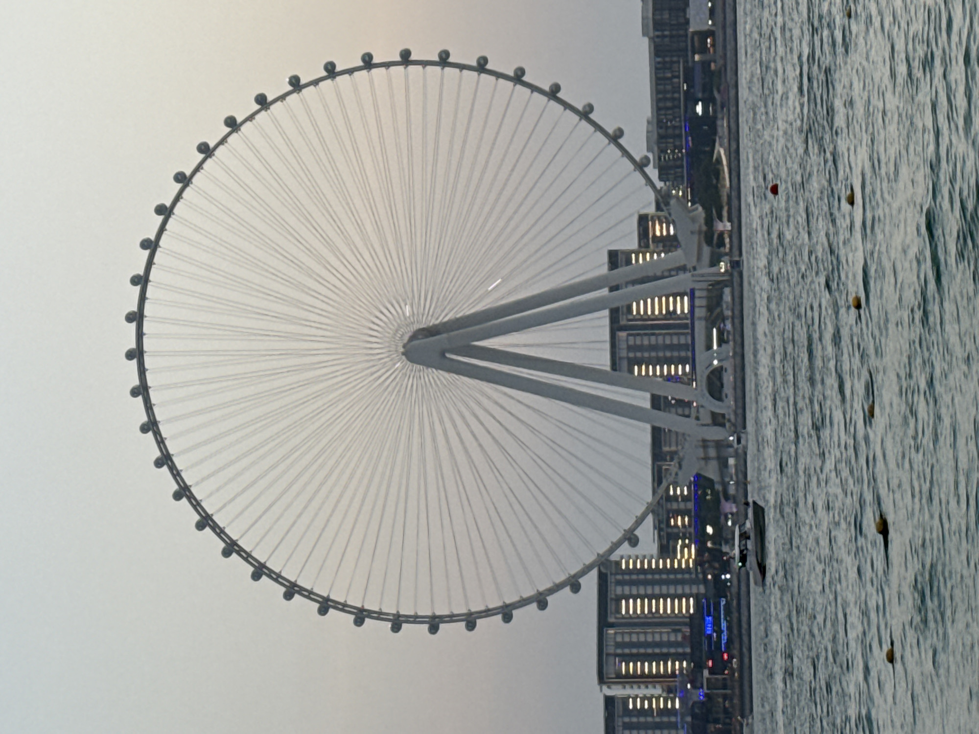 The image depicts a large Ferris wheel, a prominent object, set against a hazy sky. The Ferris wheel is circular with numerous spokes radiating from its center and pods, or cabins, attached to its outer rim. Supporting structures for the wheel are visible, extending downwards and connecting to a complex of modern buildings. These buildings are tall and rectangular, with many windows illuminated by artificial lights, suggesting an urban setting, possibly at dusk or dawn given the subdued natural light.
To the right of the Ferris wheel and buildings, a large body of water is visible, appearing choppy with whitecaps, indicating some movement or wind. Several small, yellow, spherical buoys are floating on the water's surface. The overall scene suggests a coastal or waterfront location. The lighting and the presence of illuminated buildings suggest that the time of day is either early morning or late evening. The hazy sky could indicate atmospheric conditions or pollution. No people are clearly discernible in the image. No text is visible. The location context provided is Dubai, United Arab Emirates, which aligns with the presence of such a large modern Ferris wheel and contemporary architecture.