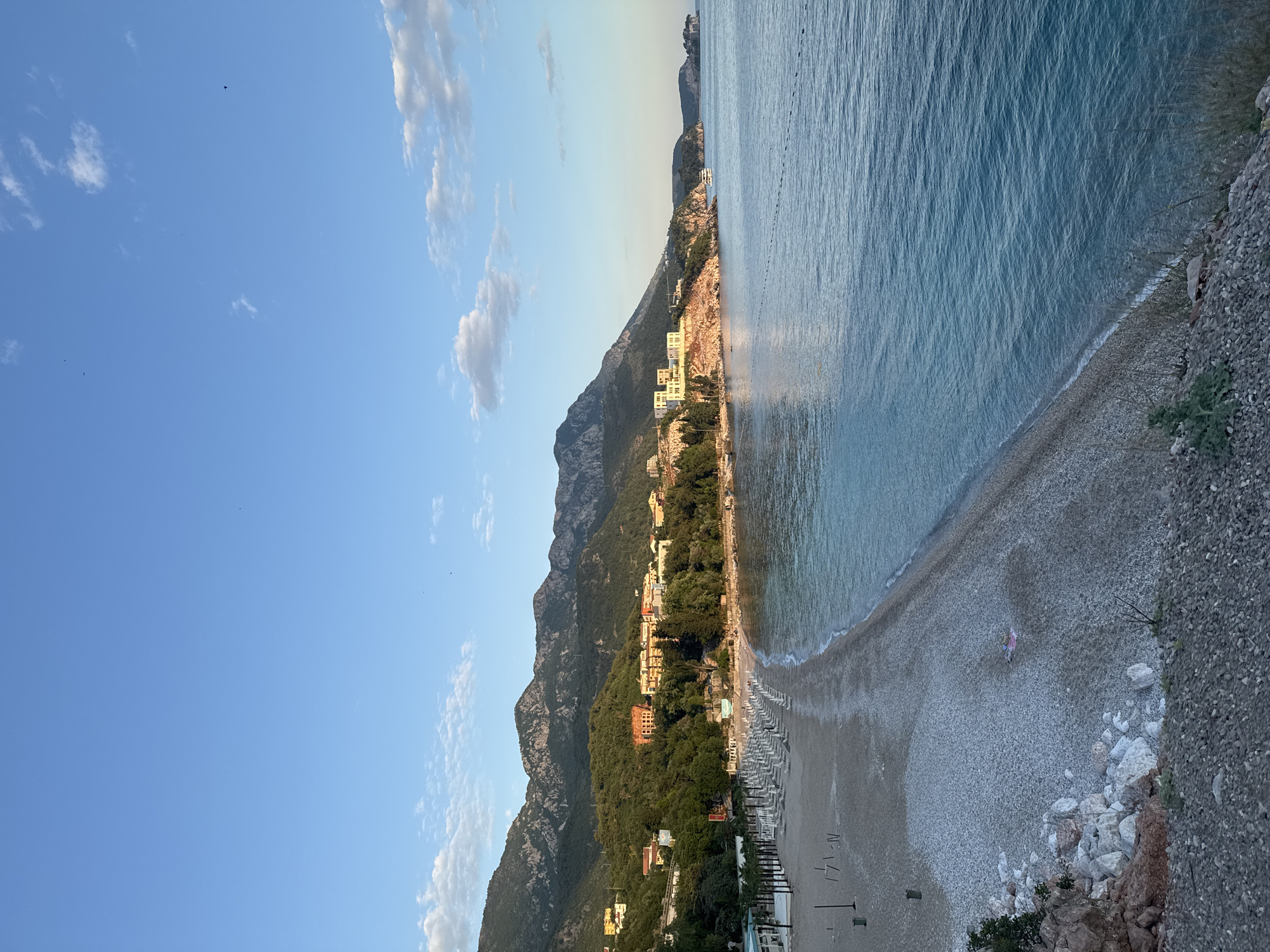 The image captures a scenic view of Kamenovo, Montenegro, showcasing a beach situated along the Adriatic coast, next to rugged mountains. The beach is mostly covered in pebbles, with rows of beach chairs, suggesting it is a tourist destination. A person in pink walks on the beach. The water is calm and clear. The mountains are densely forested, with buildings interspersed among the trees and along the coastline. The sky is blue with scattered clouds.