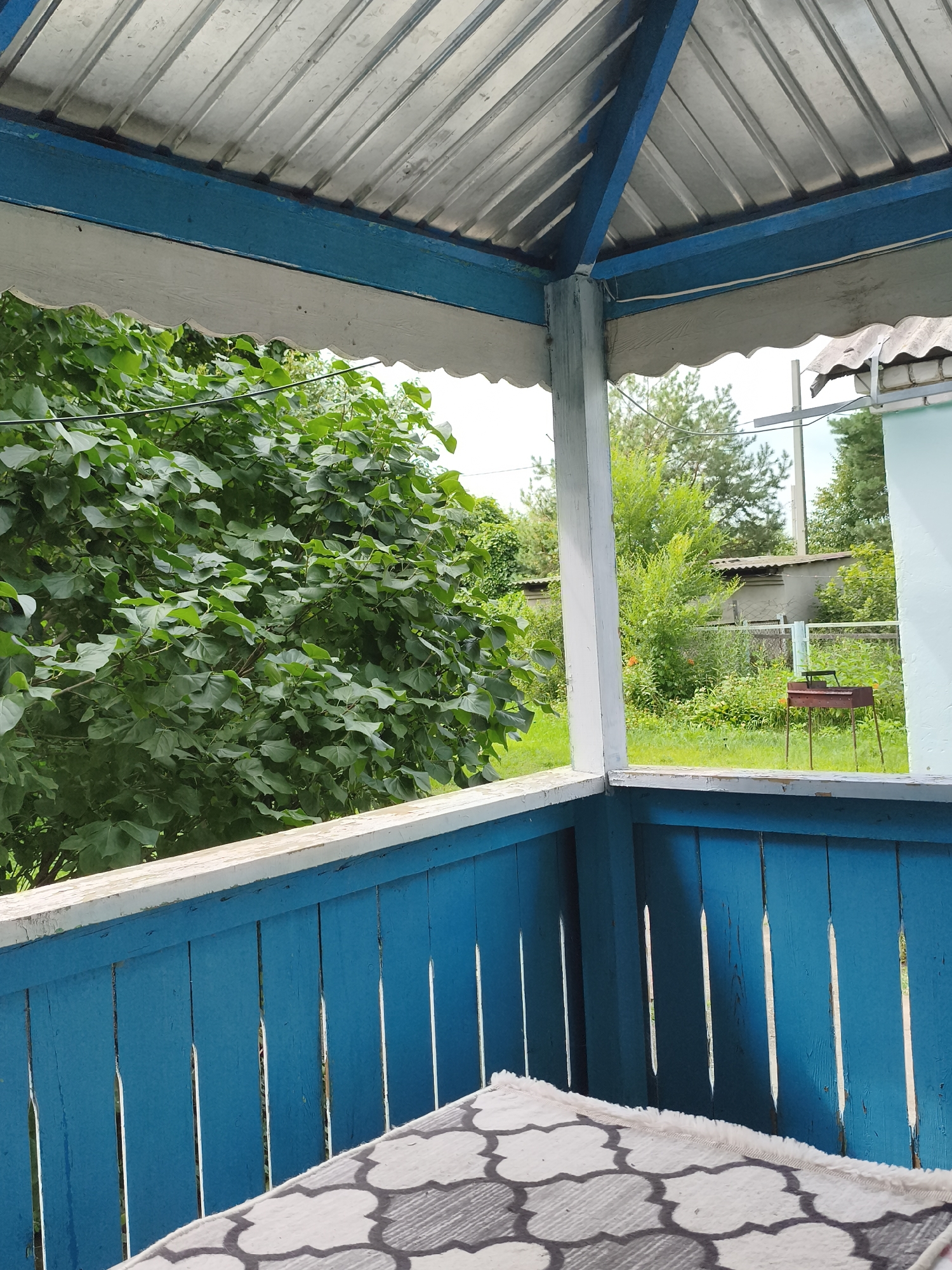 The image shows a porch with a corrugated metal roof, supported by wooden beams painted blue and white. A bright blue wooden fence runs along the edge of the porch, with a patterned rug in the foreground. Beyond the porch, there is lush green foliage and trees. In the background, a small grill is visible on a grassy area, along with a corrugated metal shed and a light blue building. The scene appears to be a residential area in Konstantinivka, Russia, likely a dacha or a backyard. There are no people or animals present in the image.