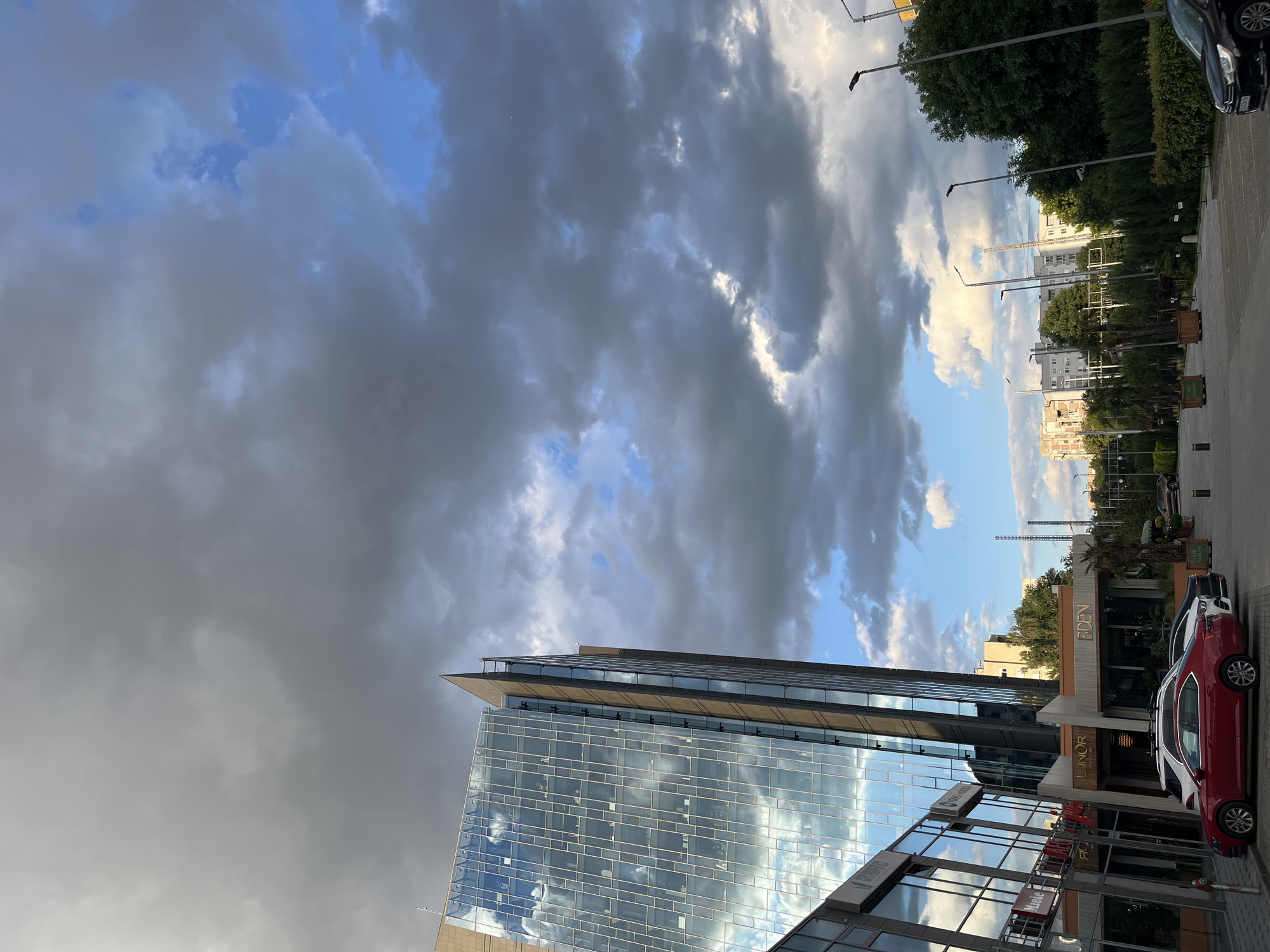 This image captures a section of a modern office building with a glass facade in Plovdiv, Bulgaria. The building, which appears to be part of a commercial or business complex, features a sharp, angled architectural element at its apex. In front of the building, a paved parking area is visible, with several cars parked, including a red sedan. The surrounding area includes other buildings, trees, and streetlights. The sky above is dramatic, with a mix of dark, cloudy formations and patches of blue sky, suggesting dynamic weather conditions. The overall scene is a snapshot of an urban environment in Plovdiv.