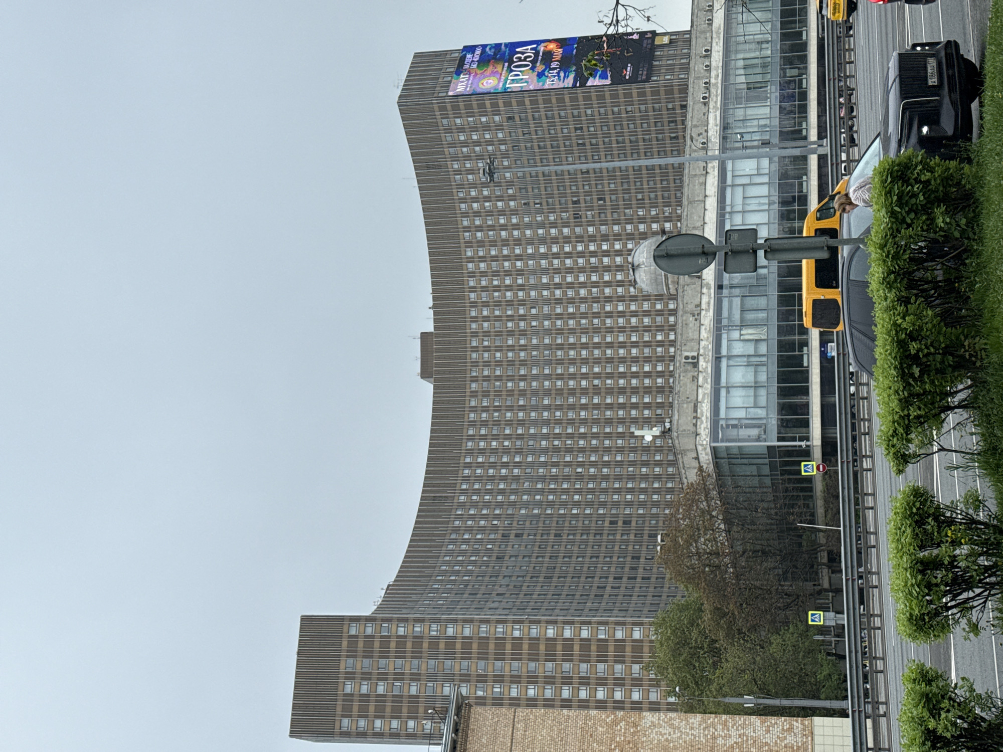 The image depicts a tall, curved building in Moscow, Russia. The building is characterized by numerous windows and a billboard at its top, displaying the word "ГРОЗА" (GROZA). A modern glass building adjoins the curved building. The scene also shows a street with vehicles, including a black SUV, a yellow van, and possibly other cars partially obscured. The street is bordered by greenery. The sky appears overcast.