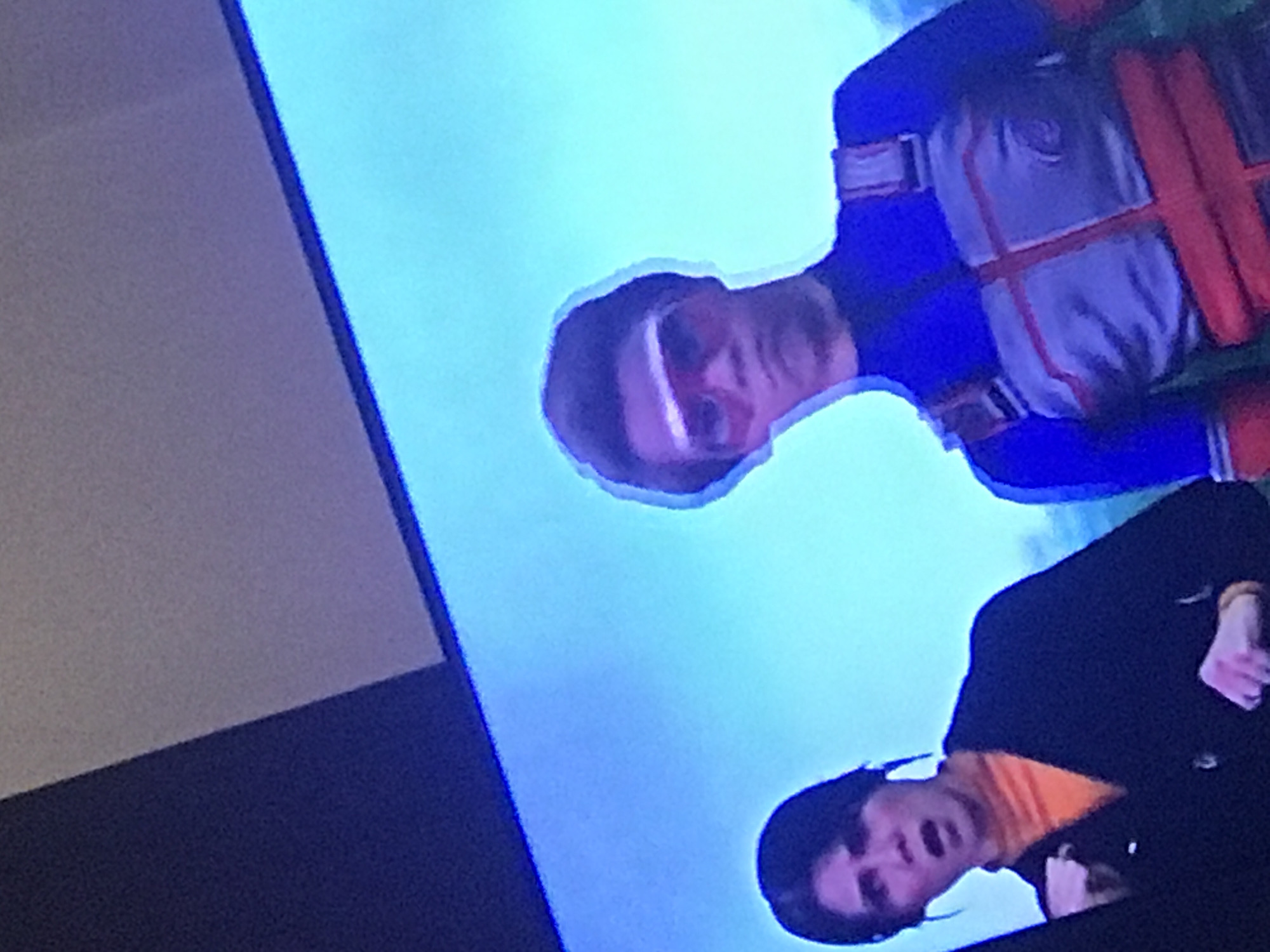 The image shows a television screen displaying two figures. One figure, in the upper part of the screen, wears a blue and white outfit with red straps. The second figure, in the lower part, wears a black jacket over an orange shirt. The image is likely taken indoors. The location is not discernible from the provided image.