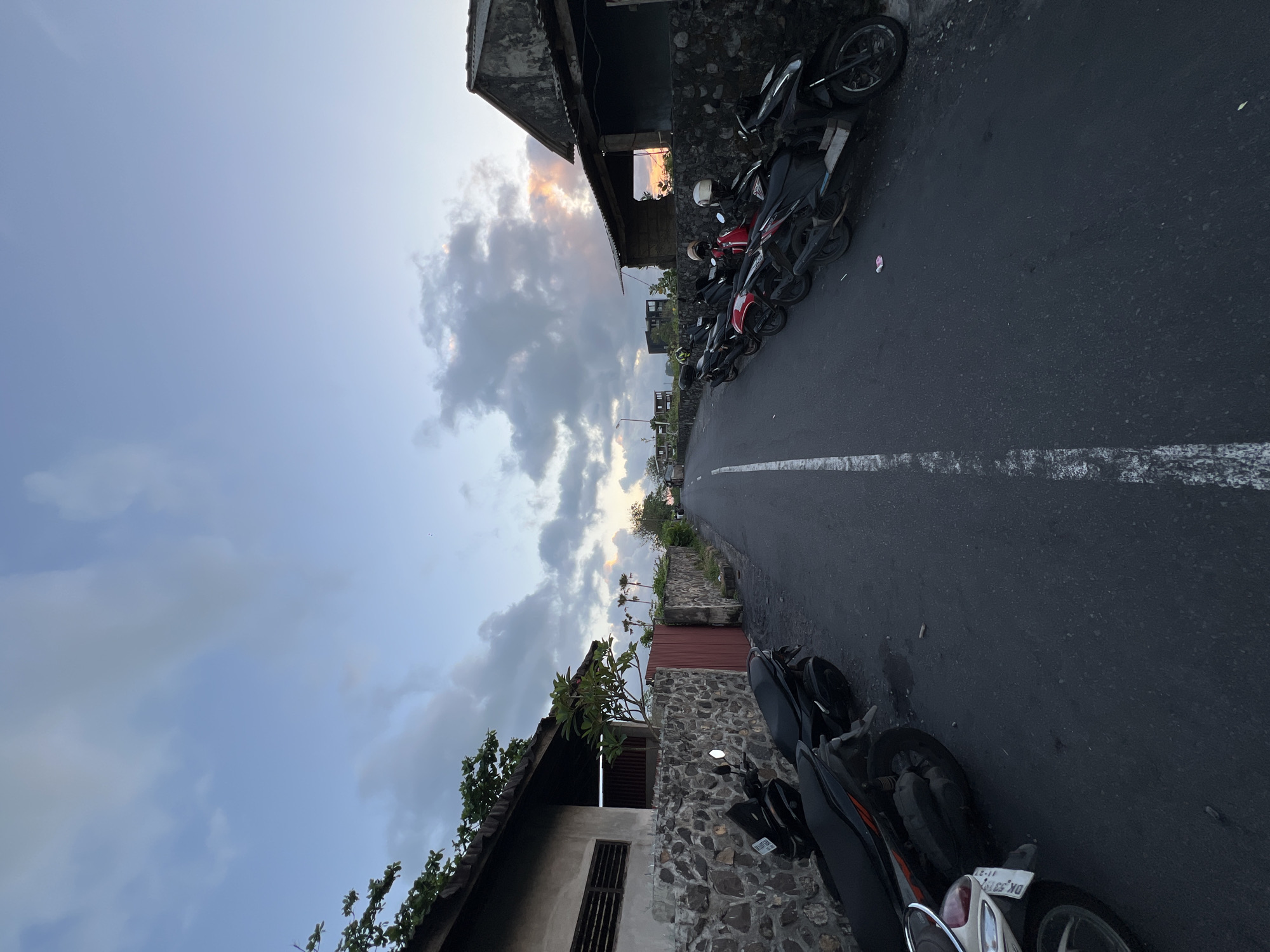 The image depicts a street scene in Unknown, Indonesia. The view is along a road with a single white line running down the center. Several motorcycles are parked along the right side of the road, adjacent to buildings with stone walls and tiled roofs. The sky is visible above, showing a mix of blue and cloudy sections with a hint of sunset colors. The angle of the photograph is rotated, making the perspective appear tilted.