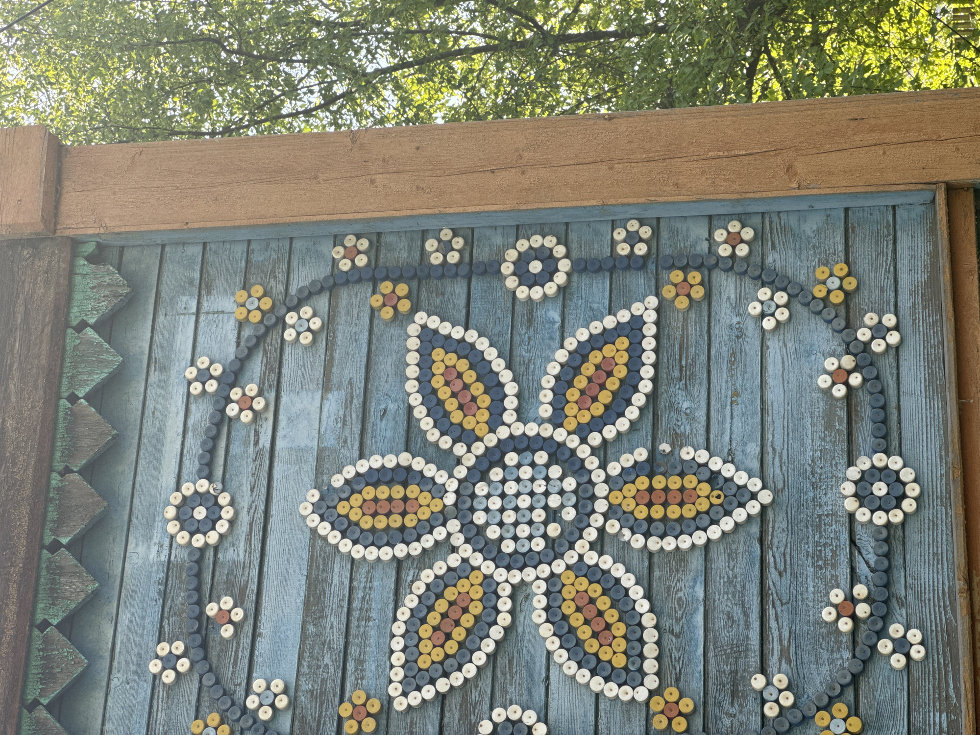 A close-up shot depicts a blue wooden fence adorned with intricate flower designs made from painted wooden spools or bottle caps. The main design is a large, stylized flower with six petals, filled with concentric circles of yellow, orange, blue, and white spools. Smaller flower motifs are scattered around, connected by lines of blue spools. The fence is topped with a wide, horizontal wooden beam, and part of a tree canopy is visible in the background. On the left side, there is a section of the fence with green and blue geometric designs. The setting suggests an outdoor location, possibly a park or yard, given the presence of the tree. The decorations on the fence give it a folk art or handcrafted appearance. The image originates from the city of Balezino, Russia.