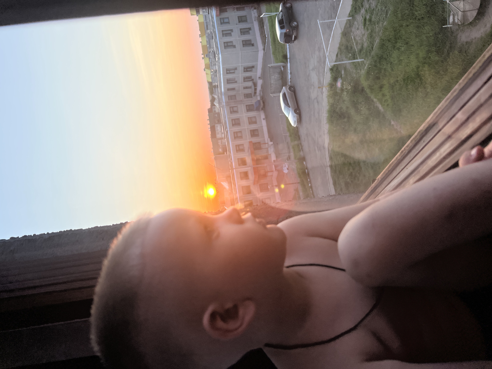 A child with a shaved head is lying down, looking out a window at a sunset. Visible outside the window are apartment buildings, parked cars, and green space, indicating a residential area in the city of Марковский, Russia. The room is dimly lit, and the child is wearing a black necklace.