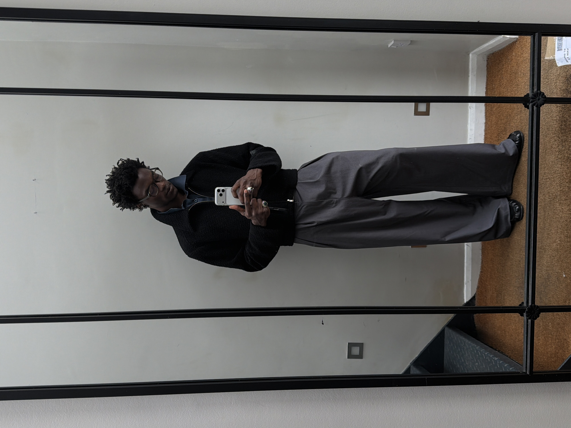 A man stands in front of a large, black-framed mirror, angled slightly to the left, holding a smartphone in his hands. He has dark skin, short curly hair, and wears glasses. He is dressed in a black sherpa jacket over a blue shirt, paired with gray trousers and black shoes. The background reflects a neutral-colored wall, an outlet, a staircase and carpet. A shipping label is affixed to the mirror near the top right corner of the frame. The location is London, UK.