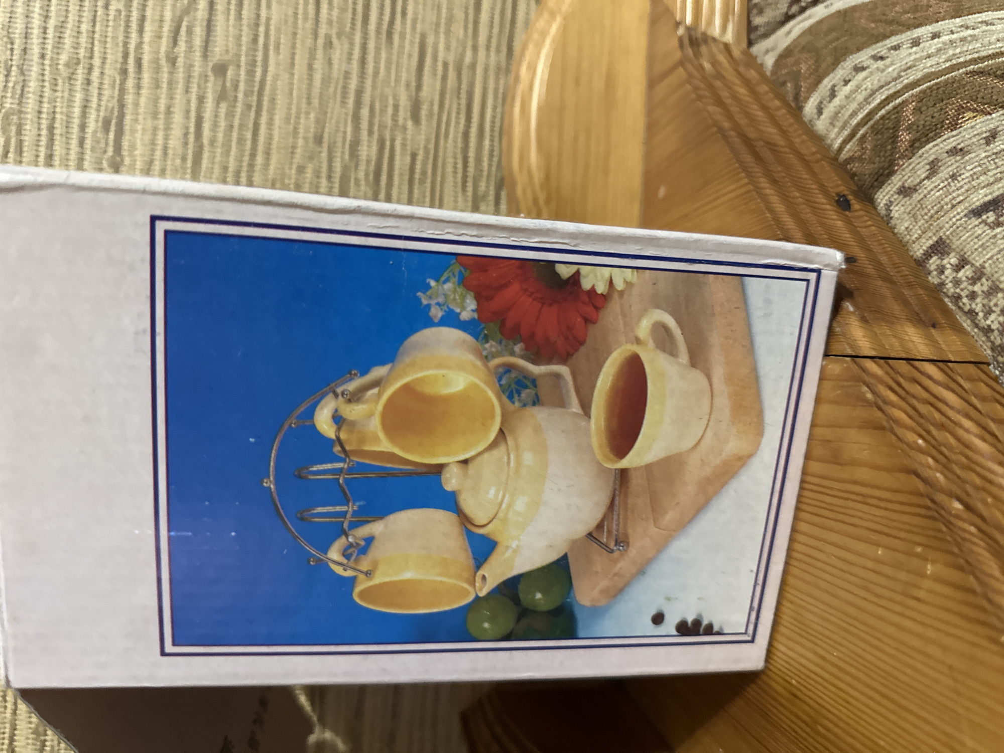 The image shows a close-up of a product box with a picture of a tea set on it. The tea set includes a teapot, several mugs on a stand, a cup of tea, some green fruits, coffee beans, and flowers. The box itself is partially visible, and it is placed on a wooden surface. The background suggests an indoor setting with wooden furniture and upholstery. There is no information in the image that indicates the location in the city of Saransk, Russia.