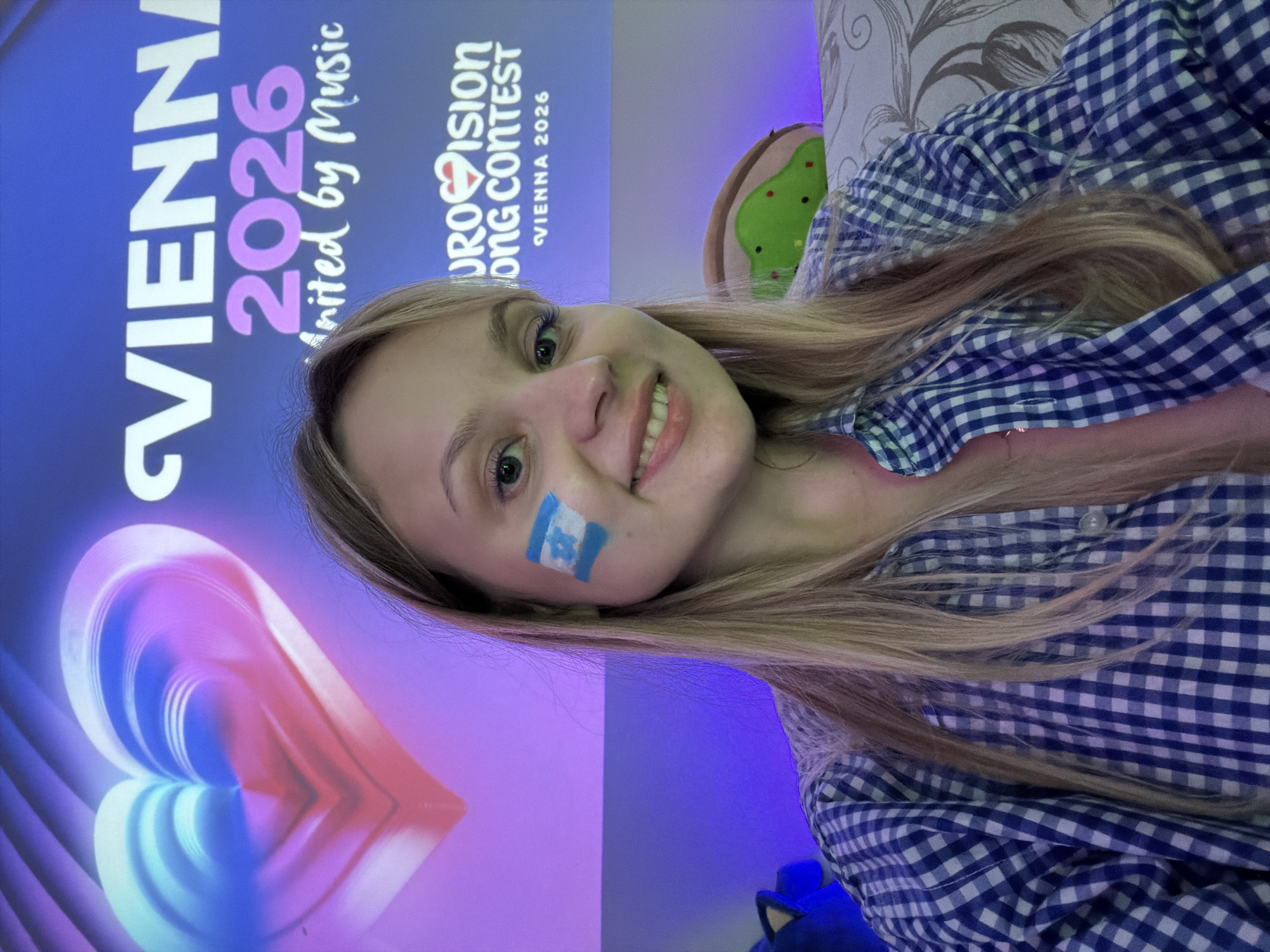 A young woman smiles at the camera with the Israeli flag painted on her cheek. She is wearing a blue and white checkered shirt. Behind her, a poster reads "Vienna 2026," along with "Eurovision Song Contest" and the phrase "United by Music." The image suggests the woman is an Eurovision Song Contest fan supporting the event in Vienna 2026. No visible indicators pinpoint the location as Bat Yam, Israel.