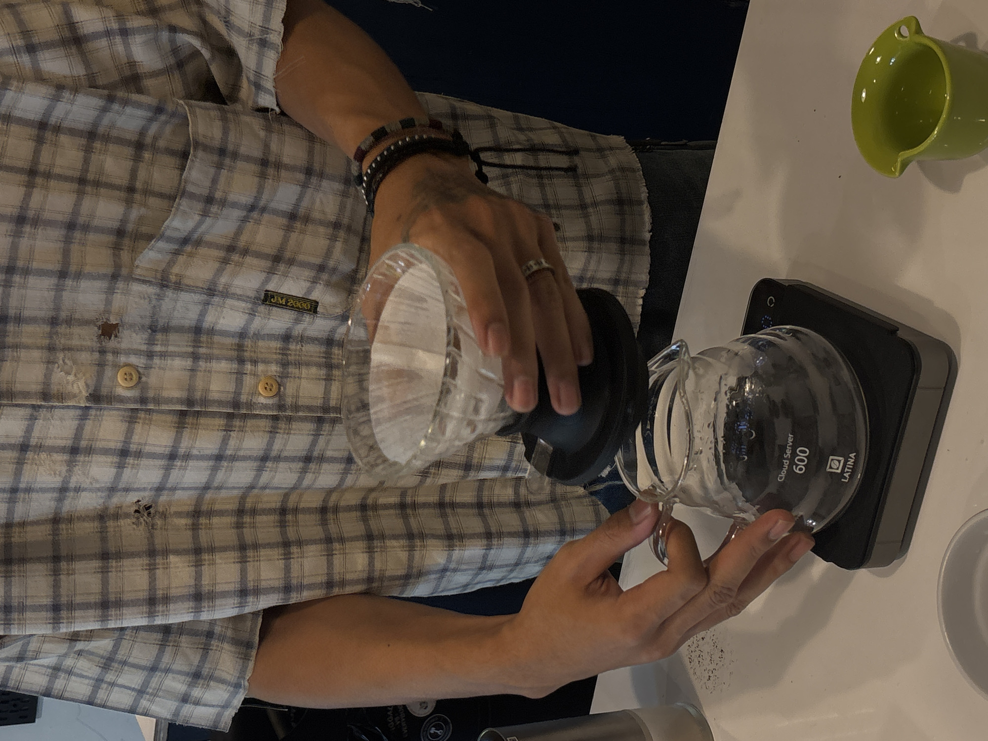 A close-up, medium shot captures a person in a plaid shirt preparing coffee. The person's hands are visible, one holding a glass carafe labeled "Cloud Server 600 LATINA" which is placed on a digital scale. The other hand holds a pour-over coffee maker with a filter and coffee grounds. A small, green ceramic cup sits to the right of the scale. The background is dark blue. The image depicts a coffee brewing process. There is no information in the image that places the scene in Semarang, Indonesia.