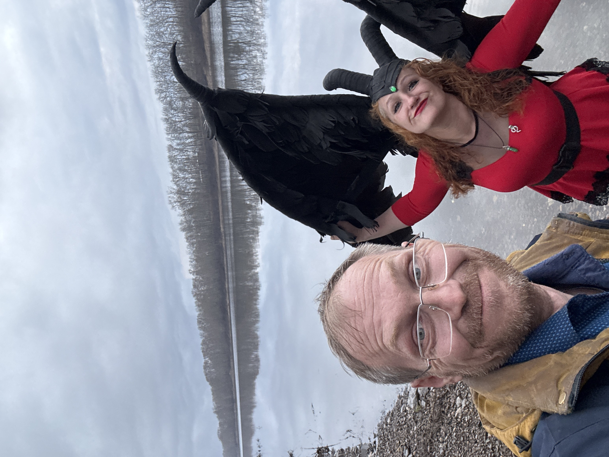 The image shows a selfie of a man and a woman with a large black swan. The man is wearing glasses and a tan jacket, with a blue shirt underneath. The woman has red hair and is wearing a red dress with black lace details, a choker, a necklace, and a horned headpiece. She is smiling and holding the swan's wing. The swan is predominantly black with a long curved neck. The background features a body of water with a line of trees on the opposite shore under an overcast sky. The ground near the people appears to be rocky. The location is near the city of Chaikovsky, Russia.