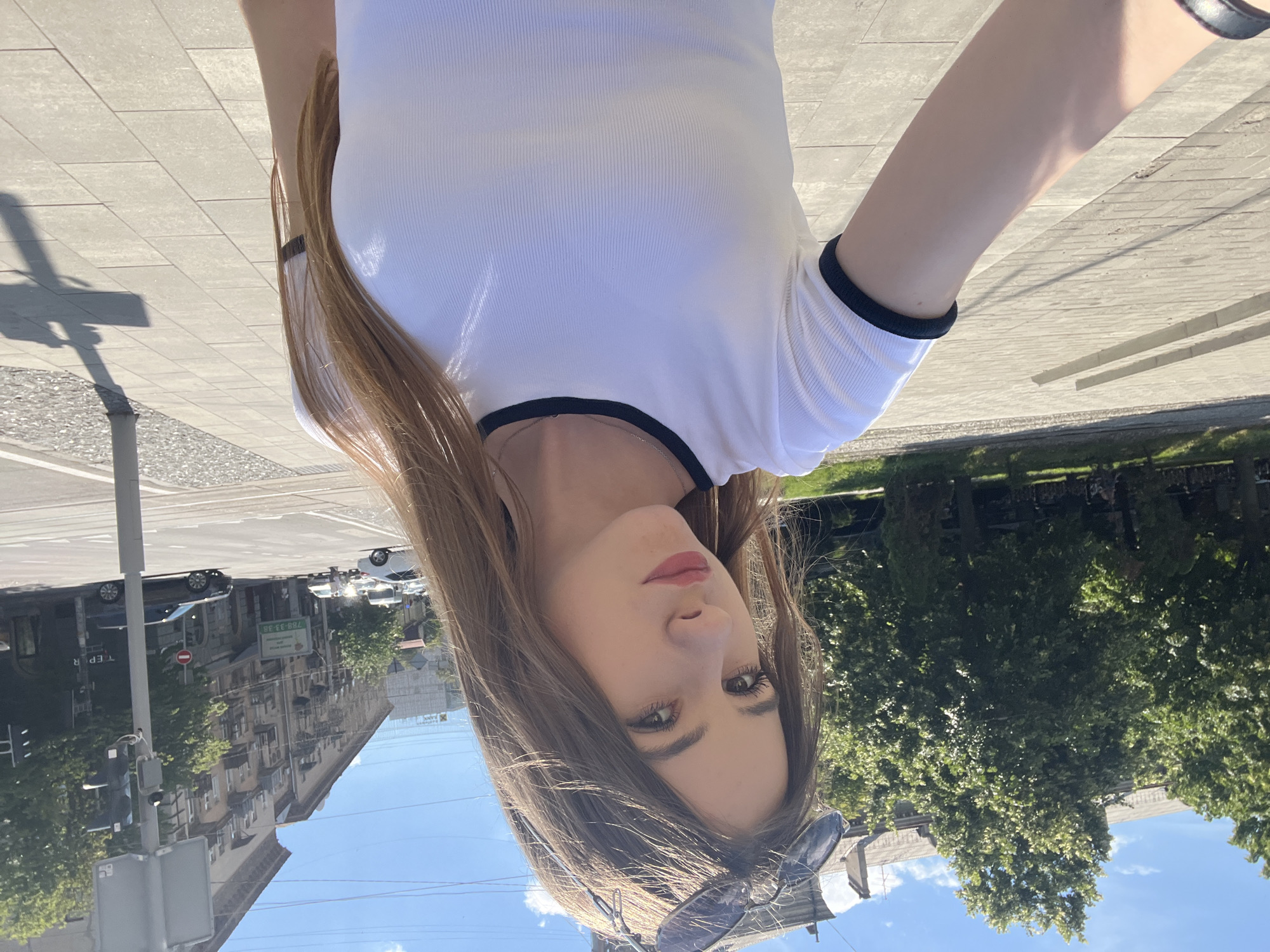 A young woman with long, light brown hair is taking a selfie outdoors on a sunny day. She is wearing a white, ribbed t-shirt with black trim around the collar and sleeves. Sunglasses are perched on top of her head. The background shows a city street with buildings, trees, and cars. To the left, there are stone steps and a sidewalk. To the right, a street light pole casts a shadow on the tiled sidewalk. The scene appears to be in Dnipro, Ukraine, suggested by the architecture and the yellow and blue banners visible in the background, which are common colors associated with Ukraine.