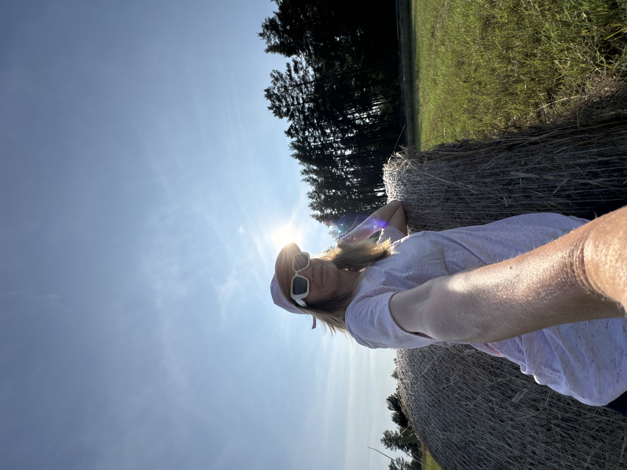 A woman is taking a selfie outdoors, with a hay bale to her left and a forest in the background. The sun is bright, creating lens flare and casting shadows. She is wearing a pink baseball cap, white sunglasses, and a light-colored t-shirt. The location is Yablonovka, Russia.