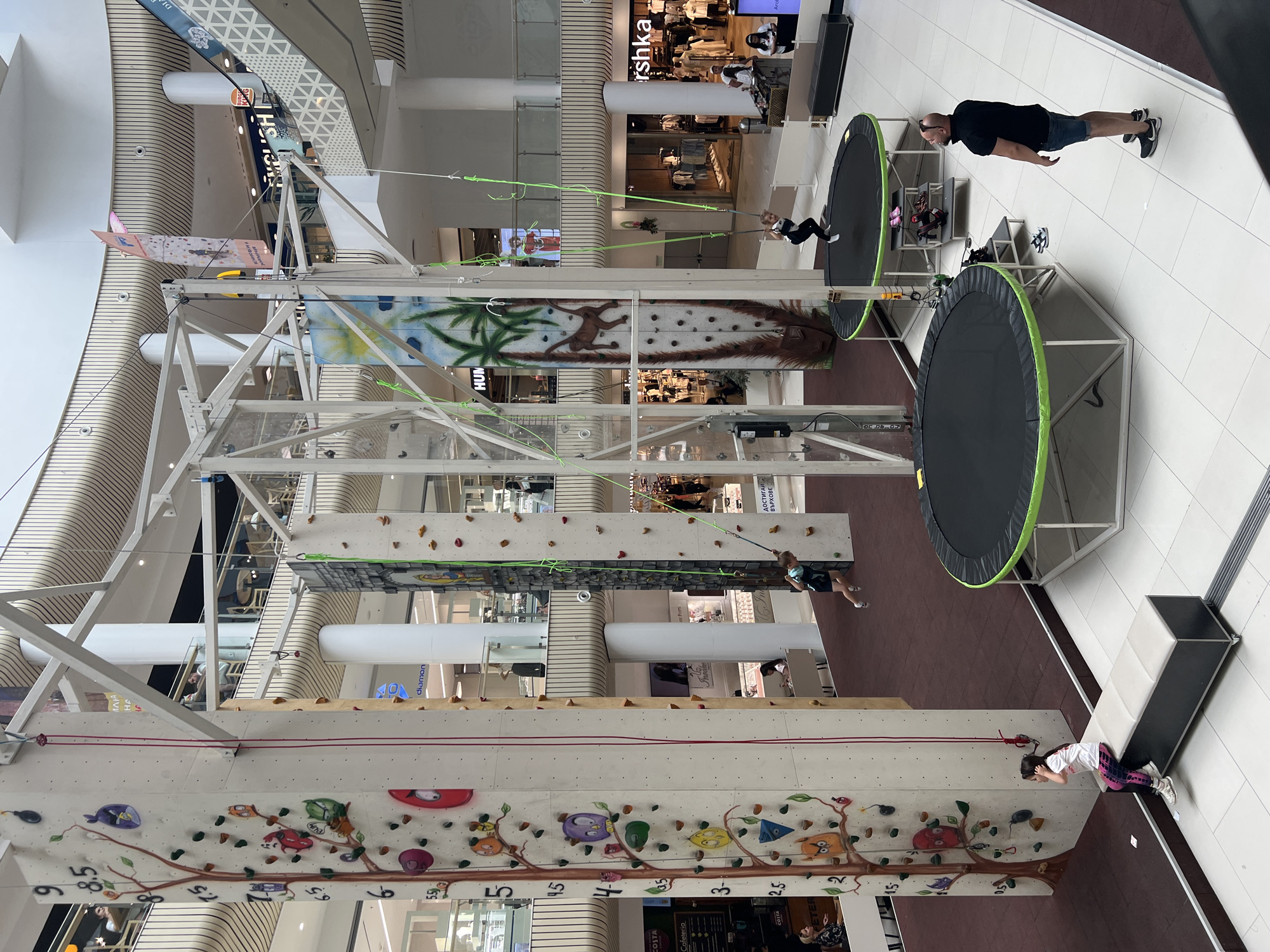 The image shows a multi-level climbing and trampoline structure located inside a shopping mall in Plovdiv, Bulgaria. The structure features a climbing wall on the bottom level, with various colorful holds and decorations. Above that are platforms with trampolines. People can be seen engaged in activities. A man stands near the upper trampoline, and a girl attempts to climb the lower wall. Mall shops and signage such as Bershka and Costa Cafeteria are visible in the background.