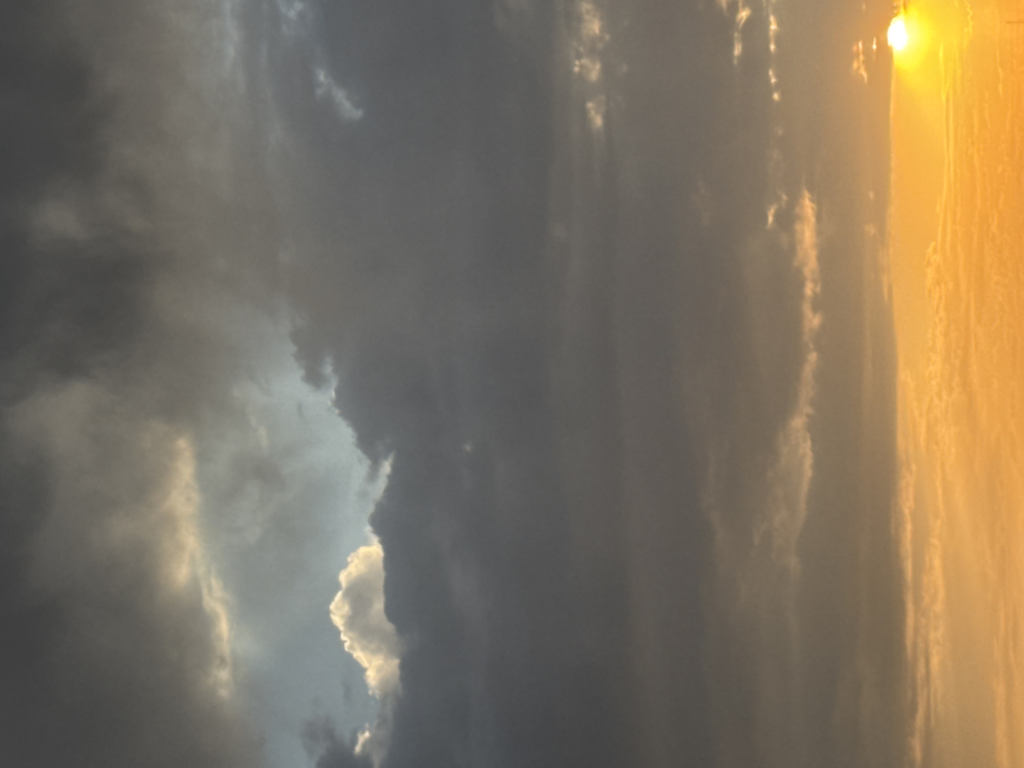 This is a vertical, medium-shot photograph of a cloudy sky at sunset. The sky is filled with dark, heavy clouds that obscure most of the sun, but a sliver of the bright orange sun is visible on the right side of the frame, low on the horizon. The lower portion of the image is a vibrant gradient of orange and yellow, indicating the setting sun. The clouds above are a mix of dark grays and lighter whites and yellows, with some areas appearing to be illuminated by the setting sun. There are no people, buildings, or any other objects visible in the image. The scene appears to be an open sky, possibly over a natural landscape or a sparsely populated area, and there is no indication that it is located in Bishkek City, Kyrgyzstan.