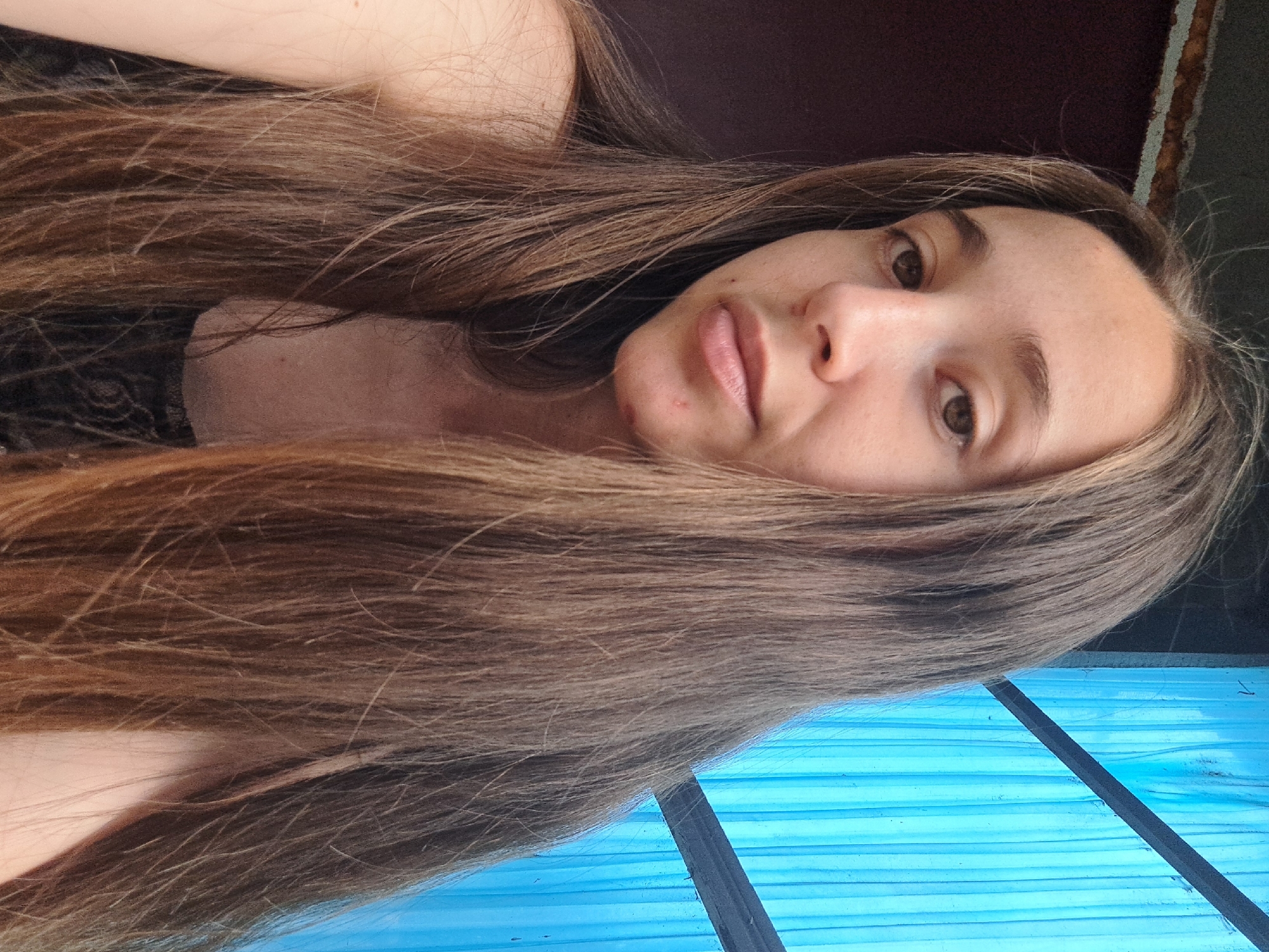 A young woman with long, brown hair is pictured in a close-up, head-and-shoulders shot. She is looking directly at the camera with a neutral expression. Her skin is fair, with some freckles visible on her nose and cheeks. She has brown eyes and natural-looking eyebrows. A few small blemishes are present on her chin. She is wearing a dark, possibly black, lace-trimmed garment. The background consists of a dark, textured surface to her left and a bright blue, corrugated plastic structure to her right, intersected by a dark horizontal bar. The lighting appears to be natural, suggesting an outdoor or well-lit indoor setting. The image was taken in Vypasne, Ukraine.