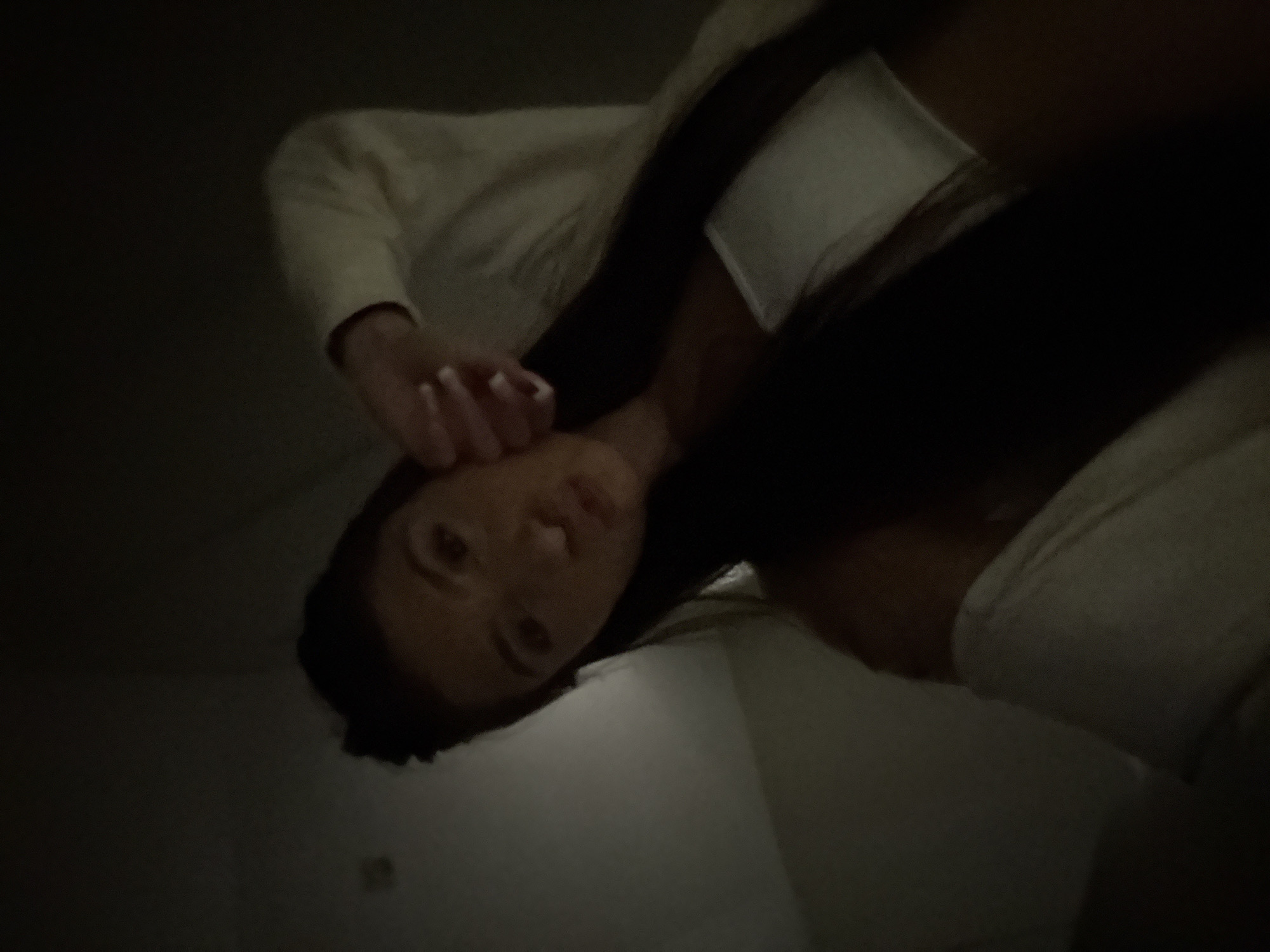 A woman is seen from a low angle, as if the viewer is lying down and looking up at her. Her face is partially illuminated, revealing dark hair, eyes, and lips. She is wearing a light-colored top. Her hand is raised near her face. The background is dark and indistinct, suggesting a dimly lit indoor space. There is no information to indicate a location in the city of Plovdiv, Bulgaria.
