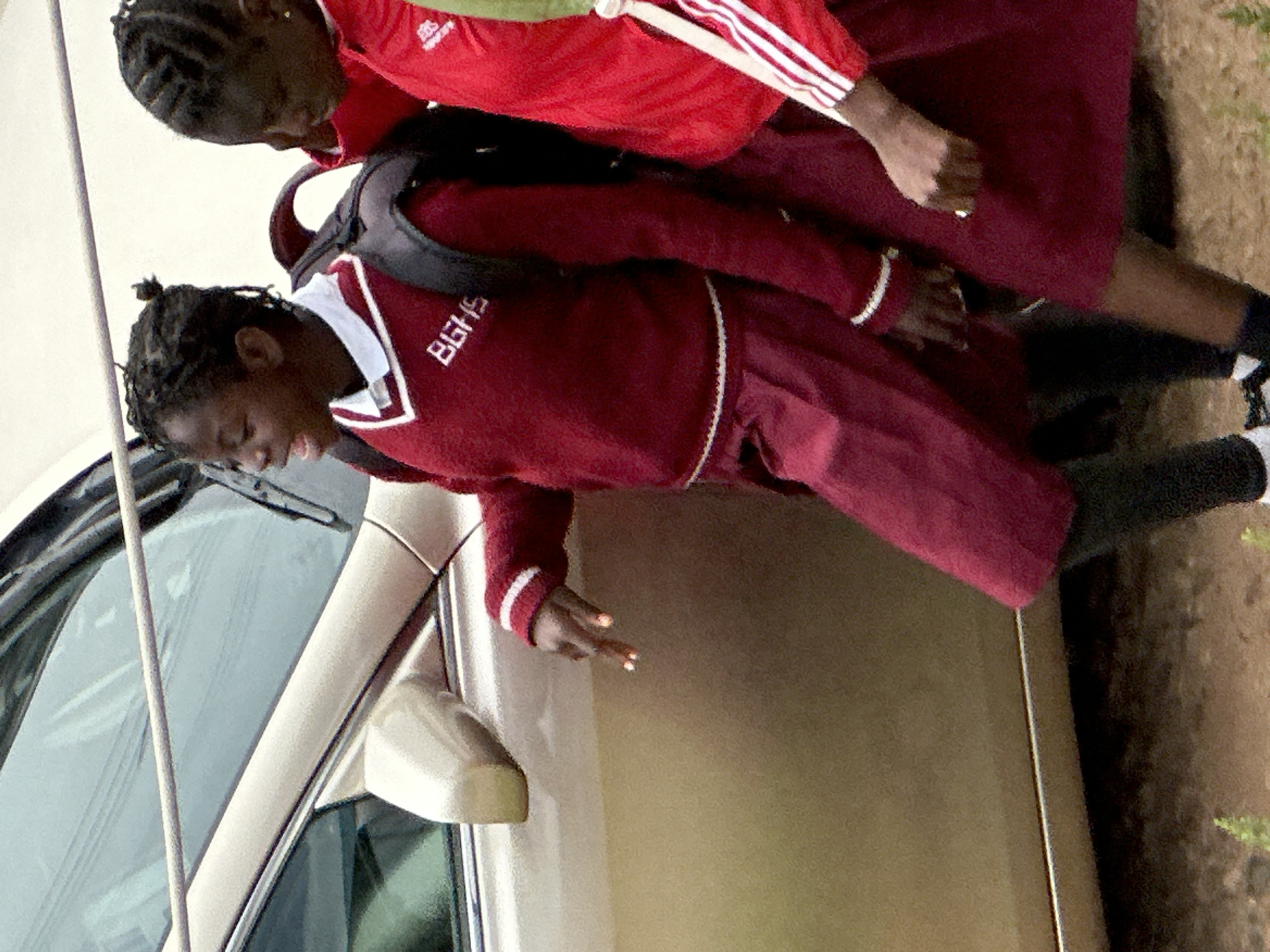 Two young girls, likely students, are interacting near the open door of a car. The girl in the foreground, wearing a maroon uniform with white trim and "BGHS" printed on it, is reaching out with her right hand. Her fingers are adorned with bright orange nail polish. She has her hair in braids, pulled back into a bun. The second girl, mostly visible from the shoulders up, wears a red tracksuit with white stripes on the sleeves and a similar maroon uniform top underneath. Her hair is also braided.
The scene appears to be outdoors, possibly a school environment given the uniforms. The car door is open, revealing the car's interior, including the dashboard and windshield. The lighting suggests it's daytime. The girls' poses imply an interaction, though the specific activity is unclear. The image is taken from a slightly elevated angle, looking down at the subjects. The setting appears to be dusty or earthy, with some greenery visible in the background. The overall impression is a candid moment captured in a casual outdoor setting.