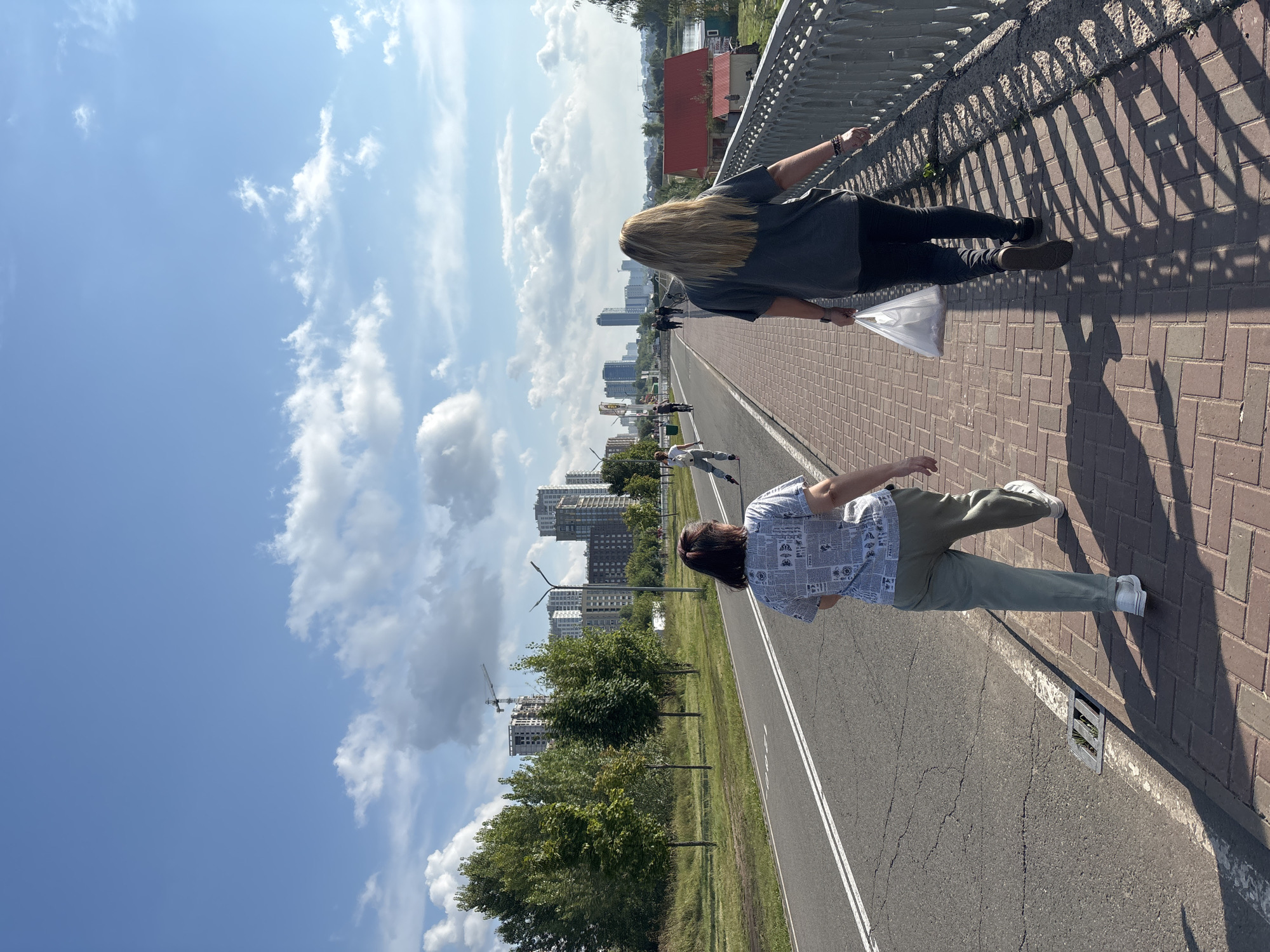 Two women are walking on a pedestrian bridge over a roadway in Naberezhnye Chelny, Russia. The woman in the foreground, on the right, has long blonde hair and is wearing a dark t-shirt and black pants. She is carrying a white plastic bag in her right hand. The woman to her left has dark hair pulled back and is wearing a patterned t-shirt and light-colored pants. She is wearing white sneakers. In the background, several other people are visible on the bridge and on rollerblades on the adjacent bike path. Beyond the bike path, there are trees and a construction site with several tall buildings and a crane. The sky is blue with scattered white clouds. The bridge has a metal railing on the right side and a brick walkway. The roadway has two lanes of traffic. The sun is casting shadows on the bridge.