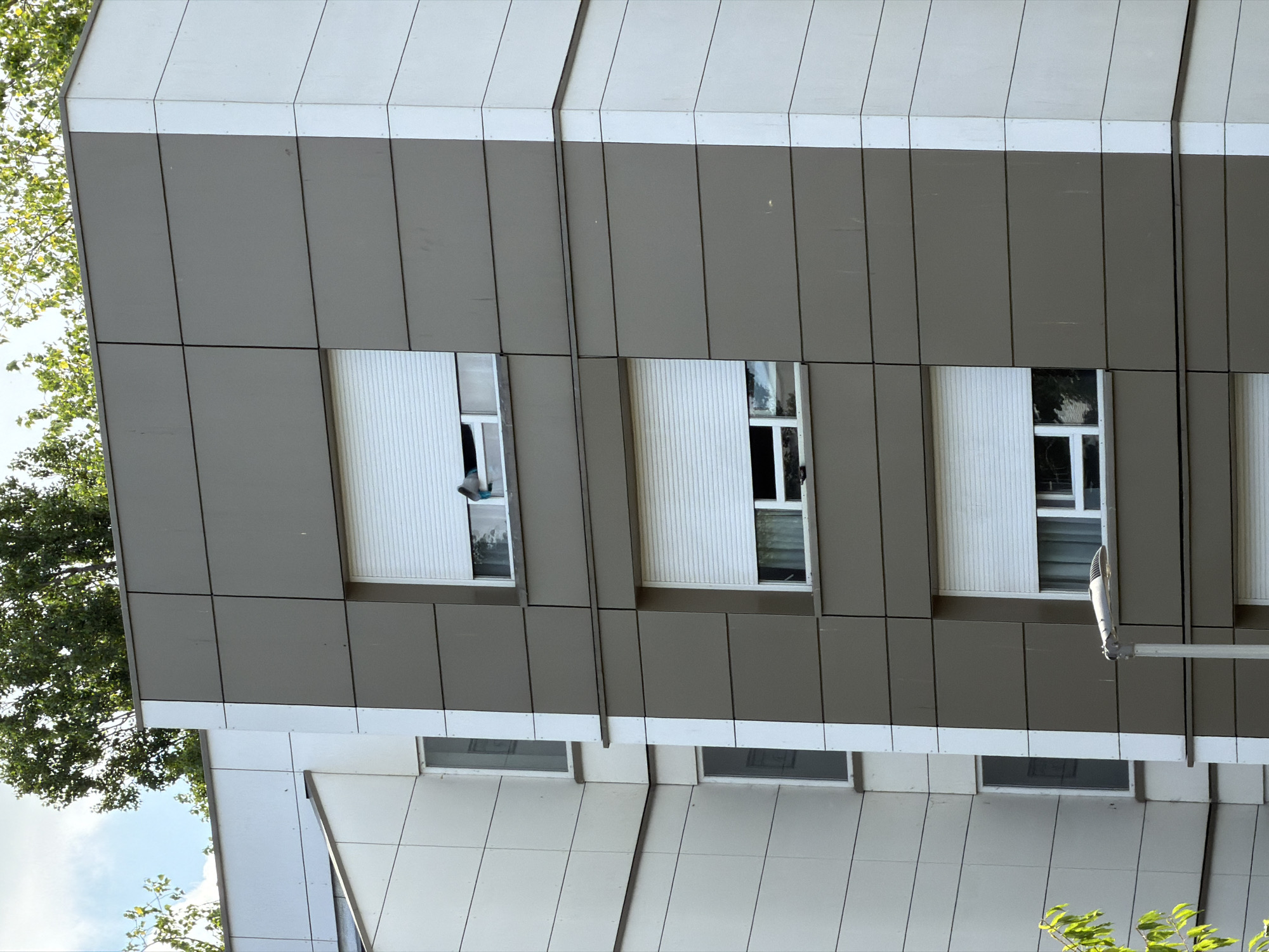 This media file shows a close-up, tilted view of a modern building's exterior. The building features a facade of alternating white and gray panels, with horizontal lines creating a segmented appearance. Several windows are visible, some with white roll-down shutters. In one of the windows, a person's head and shoulder are visible, leaning out. Trees with green foliage are seen on the left side of the frame, suggesting an outdoor setting. The sky, partially visible, is blue with some clouds, indicating daytime. A street lamp is mounted on the building's facade on the right. The lower part of the building displays more windows, some with dark interiors. The overall impression is of a contemporary residential or office building. The location is identified as Évry-Courcouronnes, France. No text is visible in the image.