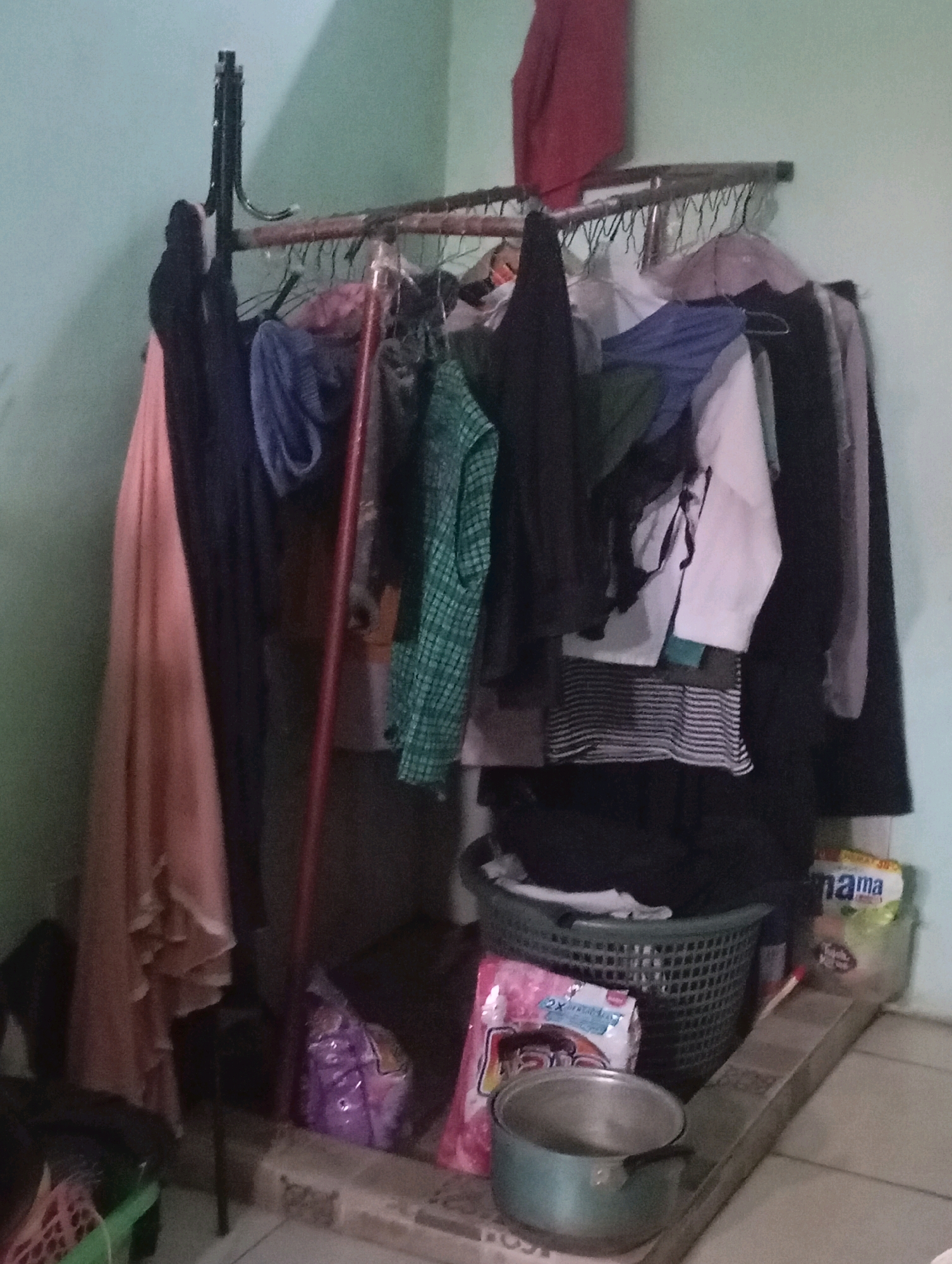 An interior scene in a dimly lit room, likely a dwelling, in Babelan, Indonesia, features a clothes rack laden with various garments. The rack is made of metal poles and a wire mesh. Hanging from the rack are shirts, dresses, and possibly other clothing items in a variety of colors including pink, black, blue, green, and white. Some items are folded, while others are draped. To the right of the clothes rack, a dark green plastic laundry basket sits on the floor, filled with clothes. In front of the basket is a pink plastic bag, and to the left of that, a light blue metal pot with a handle. Behind the laundry basket and to its right, a white plastic bag with the word "mama" printed on it is visible. The floor appears to be tiled. The walls of the room are painted a light blue-green. The overall impression is of a functional, everyday space with laundry and clothing storage.