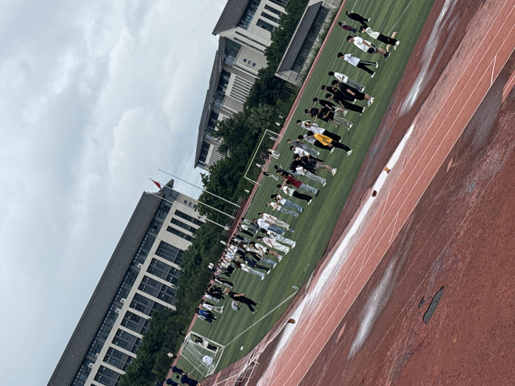 The image shows a group of students on a green sports field, likely a school campus, with large, modern buildings in the background. The students are dressed in casual attire, a mix of t-shirts, jeans, and athletic wear, and appear to be organized in rows, suggesting a planned activity such as a physical education class or assembly. Some students are looking towards the front, while others are interacting with their peers. The field itself appears to be a synthetic turf with a running track made of red material in the foreground. The track shows signs of wear and tear, with some damp patches and cracks visible. The sky is overcast, indicating a cloudy day. The architecture of the buildings suggests an institutional setting. Based on the visual cues, this scene is likely located in Jiangjin District, Chongqing, China, a region known for its educational institutions and modern infrastructure, consistent with the buildings depicted.