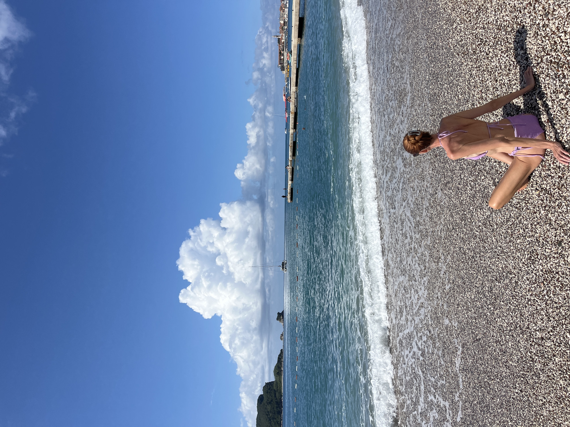 The image, when viewed rotated to its correct orientation, depicts a bright, sunny day on a pebble beach in Budva, Montenegro. In the foreground on the right, a woman with reddish-brown hair, wearing a lavender two-piece bikini, is captured from behind. She is kneeling or crouching on the small-stone beach, with her arms extended forward and hands resting on the pebbles, appearing to be in a relaxed pose.
To her left, the clear, turquoise-blue sea gently meets the shore, with delicate white foam from small waves washing over the smooth pebbles. A long pier or breakwater extends across the midground, with several small boats and yachts anchored or sailing near it. In the distance, along the coastline, parts of a town with traditional buildings and red-tiled roofs are visible. Further to the left, verdant hills or mountains rise from the sea, framing the horizon.
The sky is a brilliant, deep blue, occupying a significant portion of the image. Prominent, large, fluffy white cumulus clouds are scattered across the sky, particularly a very expansive cloud formation dominating the left side. The lighting suggests mid-day, with strong sunlight illuminating the scene. The overall atmosphere is one of a peaceful, pleasant day at the beach. There is no visible text in the image.