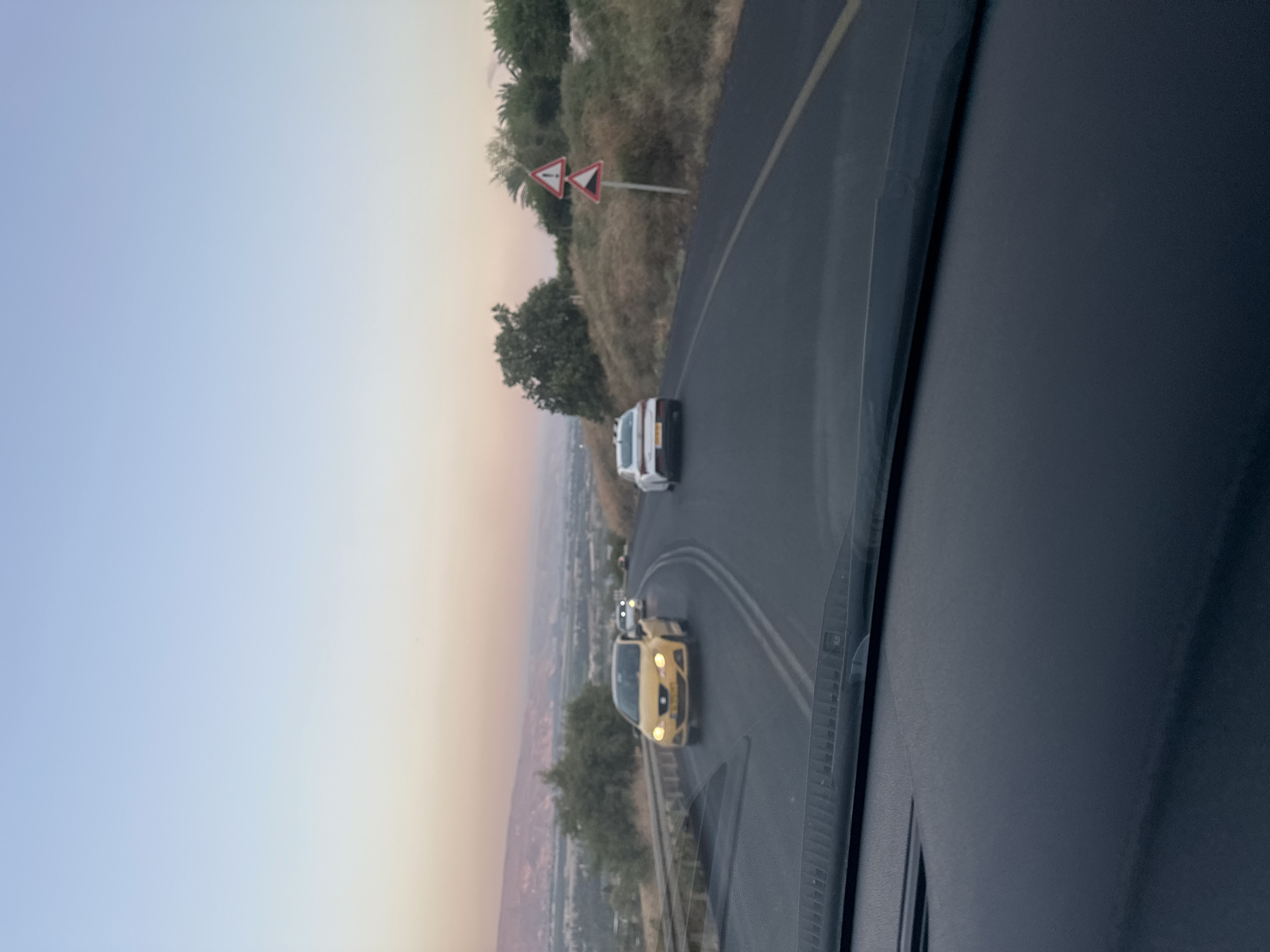 The image captures a view from inside a vehicle traveling on a road in the Emek HaYarden Regional Council, Israel. The road curves to the right, with a yellow car in the foreground leading a white car and other vehicles in the distance. The landscape features rolling hills covered in dry vegetation and scattered trees, with a distant valley and mountains under a pale sky at dusk. Two triangular road signs are visible on the right side of the road: one depicting an exclamation mark and another showing a steep descent. The dashboard of the vehicle is visible at the bottom of the frame.