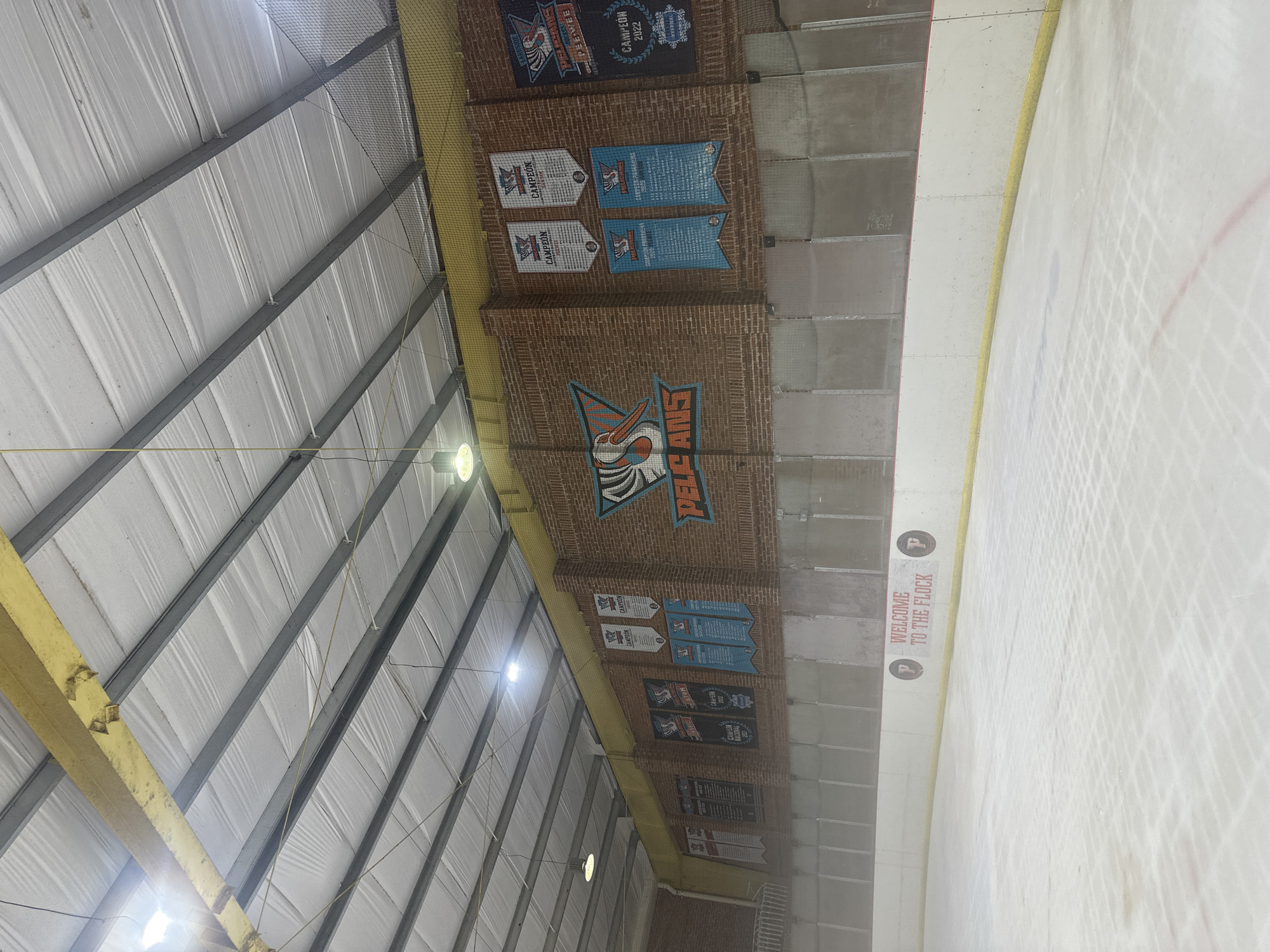 The image depicts the interior of an ice rink, likely located in Juriquilla, Mexico. The rink features an ice surface with red and white markings. Along the wall, a series of banners are displayed, celebrating championships. The banners are predominantly white and blue and feature the logo of a pelican. One large banner prominently displays the words "PELICANS" alongside a stylized pelican graphic. Another sign on the rink's barrier reads "WELCOME TO THE FLOCK." The ceiling of the rink is constructed with metal beams and white insulation panels, with lighting fixtures suspended from above. The walls are made of brick and paneling. The overall impression is that of a sports facility dedicated to ice hockey or figure skating.