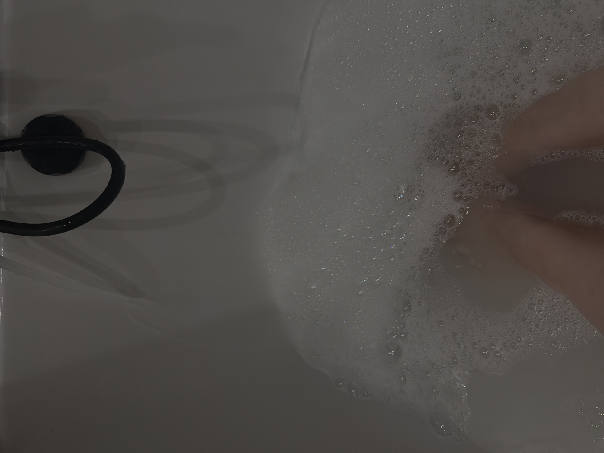 A high-angle, indoor shot captures a partial view of a white bathtub filled with soapy water and bubbles. A black, curved towel rack is attached to the side of the tub, and a person's legs are submerged within the bubbles. No specific information indicates that the image was taken in Reshetnikovo, Russia.