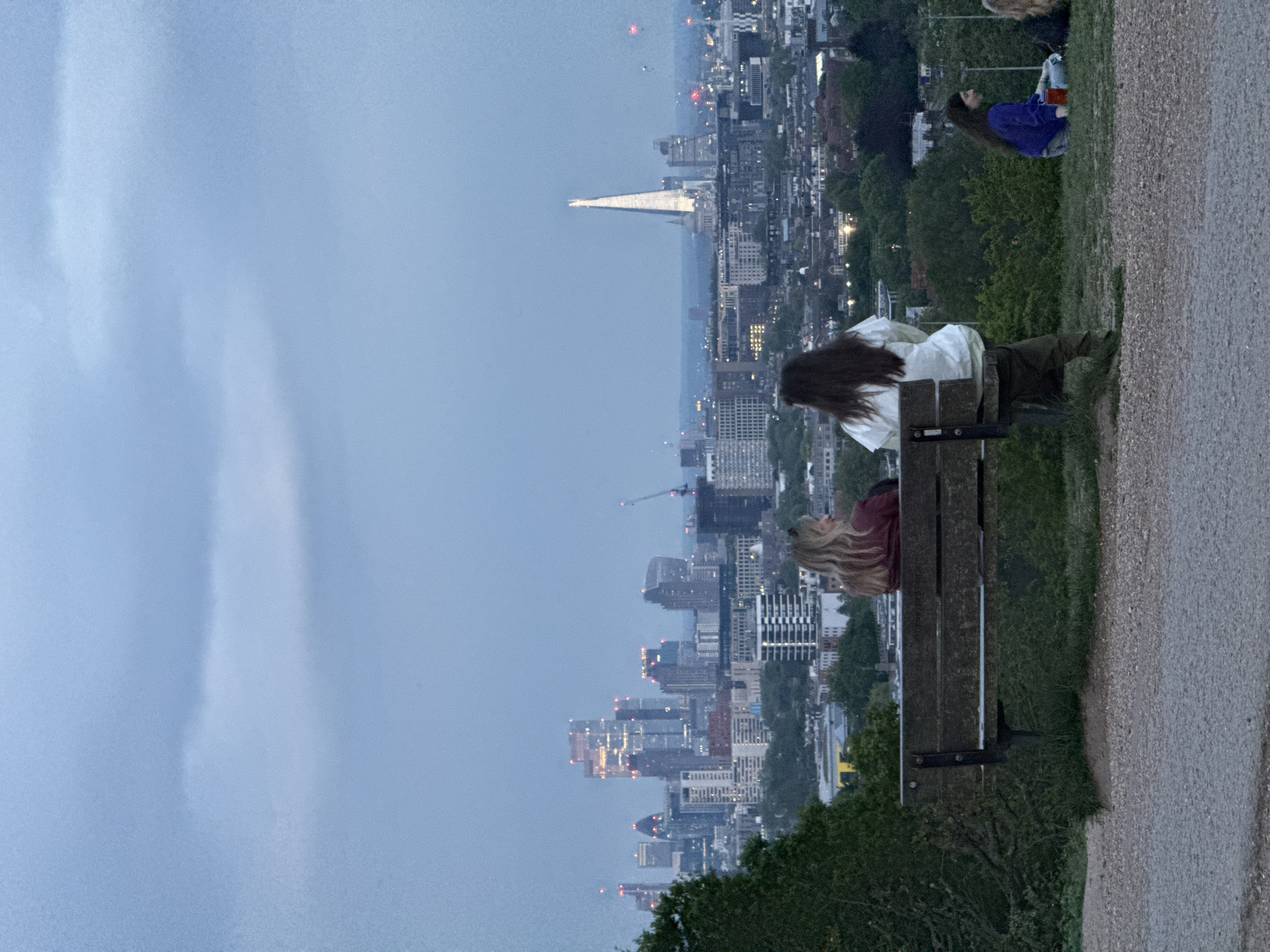 Two women are sitting on a park bench overlooking the city of London. One woman with long brown hair is wearing a white shirt and dark pants. The other woman with long blonde hair is wearing a maroon shirt. A third woman is reclining in the grass nearby, wearing a blue shirt. The bench is located on a slight hill. The city skyline is visible in the background with the Shard in the distance. The sky is overcast.