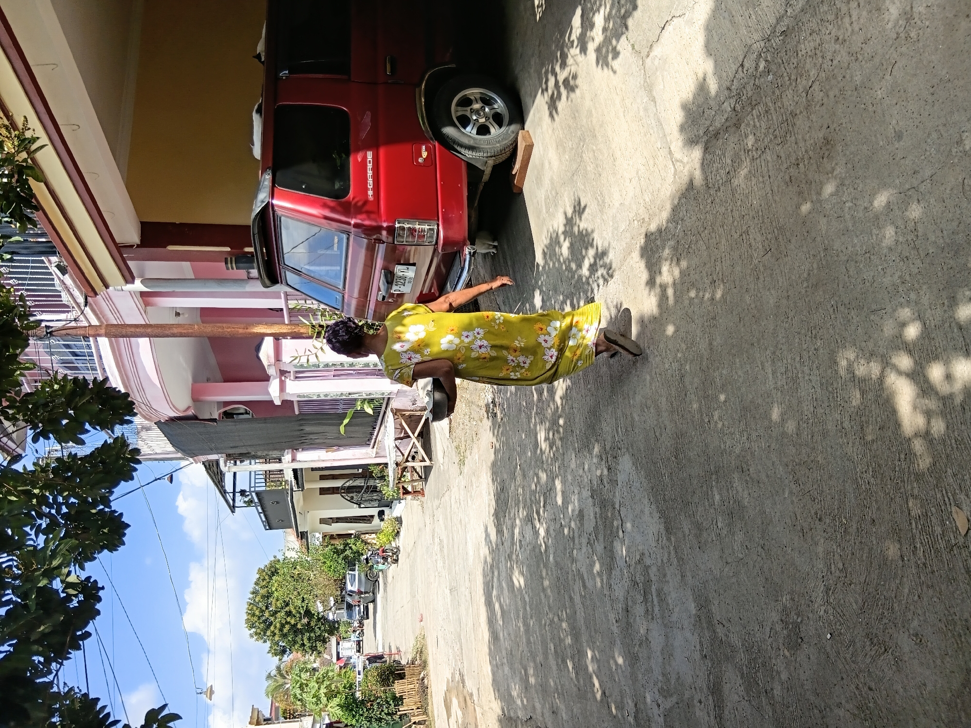A woman wearing a yellow floral dress walks away from the camera on a paved street. She is carrying a dark, round bowl in her left hand. To her right is a red SUV parked in a driveway. The street is lined with houses and trees. The sky is blue with some white clouds. The location is identified as Tanggul Wetan, Indonesia.